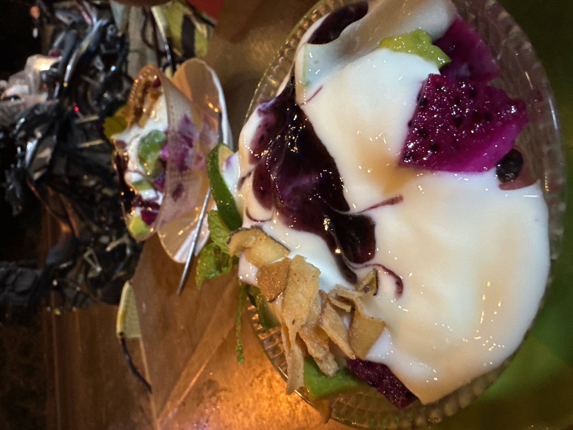 This image, likely captured in Mù Cang Chải, Vietnam, displays a vibrant dessert served in a clear glass bowl, viewed from an overhead perspective. The dessert features a base of creamy white substance, possibly yogurt or a similar dairy product. It is adorned with chunks of deep purple dragon fruit and smaller pieces of green fruit, which could be kiwi or green dragon fruit. A dark purple sauce or syrup is drizzled across the white cream, creating an appealing swirl. The dish is garnished with thin, golden-brown crispy flakes, possibly fried coconut or a similar topping, and fresh green mint leaves.
In the background, another similar dessert bowl is visible, out of focus. Both bowls rest on a warm-toned wooden surface. The lighting is dim and artificial, casting warm glows and reflections on the dessert's surface, suggesting an evening or nighttime setting indoors. No people are explicitly visible, though blurred elements in the far background might indicate their presence. No text is discernible in the image.