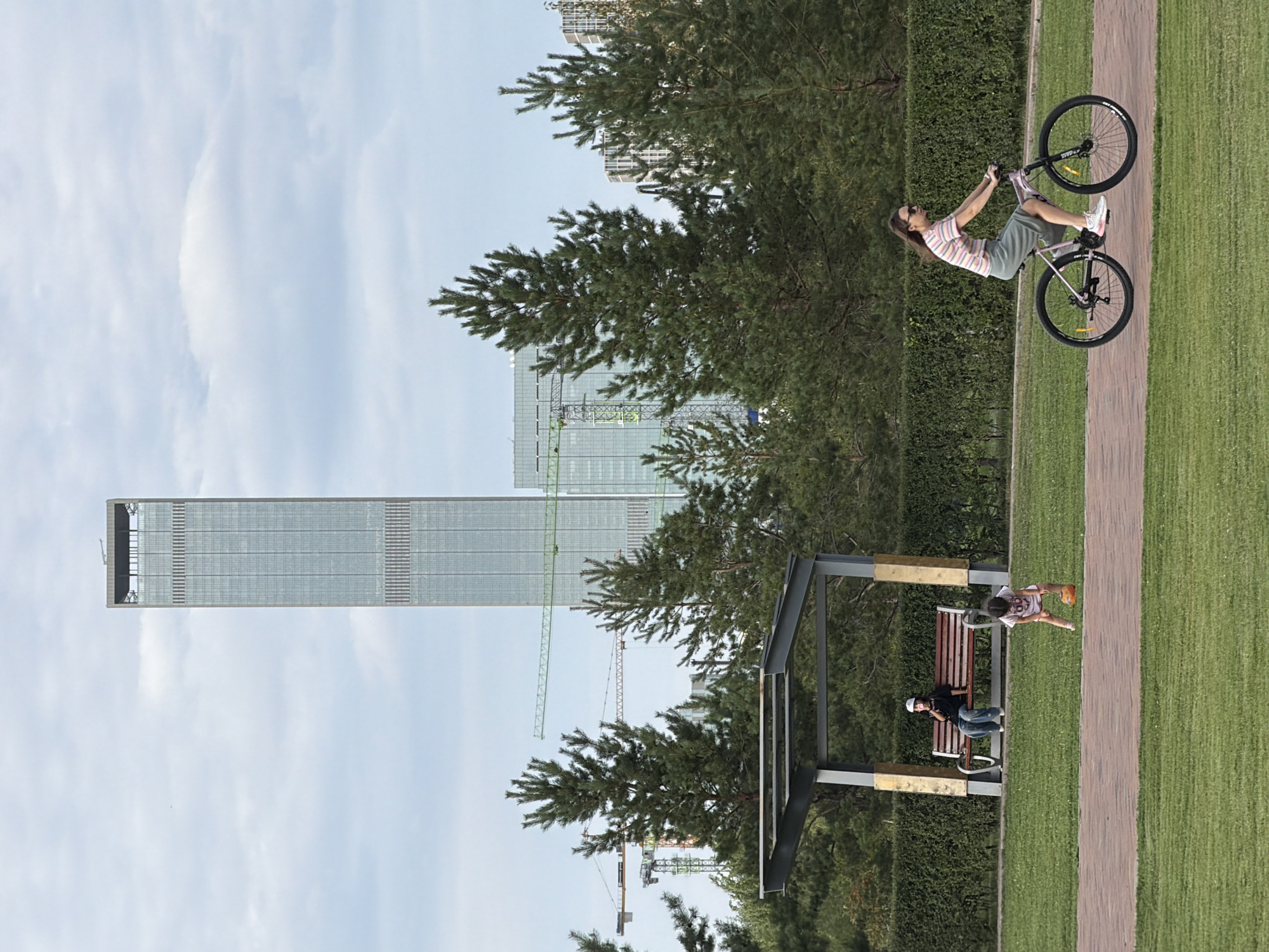 In Astana, Kazakhstan, a scene unfolds in a park with a prominent skyscraper in the background, suggesting an urban setting. On the left, a person sits on a park bench under a covered shelter, wearing a cap and jeans. To the right, a young child is playing with a ball on the grass. Further to the right, a woman is riding a bicycle on a paved path. The environment includes well-maintained green grass, a pathway, a hedge, and pine trees, with construction cranes visible behind the trees, indicating ongoing development. The sky is overcast with clouds.