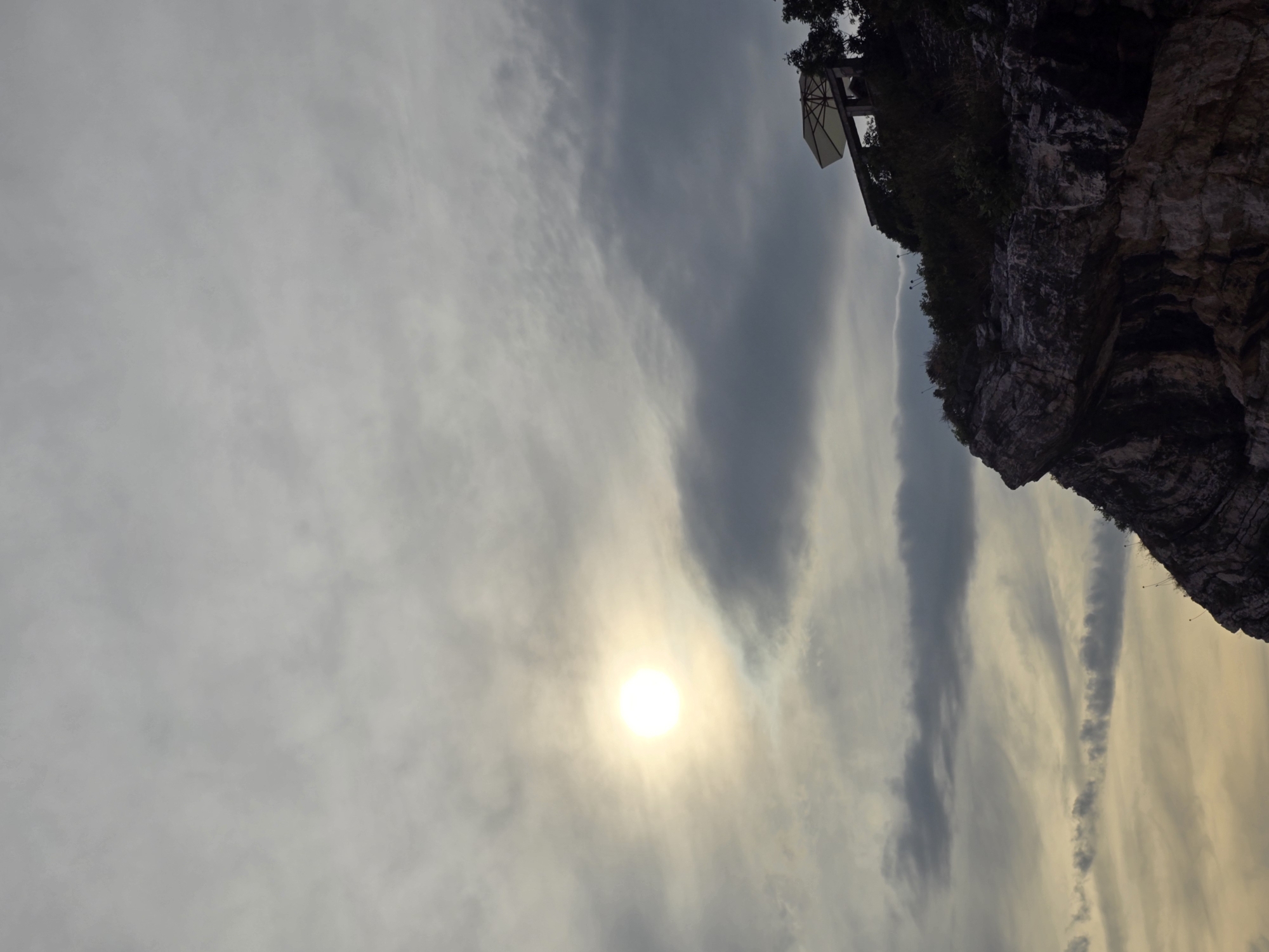 The image depicts a cliff face in Mirište, Montenegro, with a structure and umbrella on top. The sun is visible in the sky, partially obscured by clouds. The cliff is rocky and covered in vegetation. The overall scene is overcast and appears to be a natural landscape with a man-made structure on the edge.