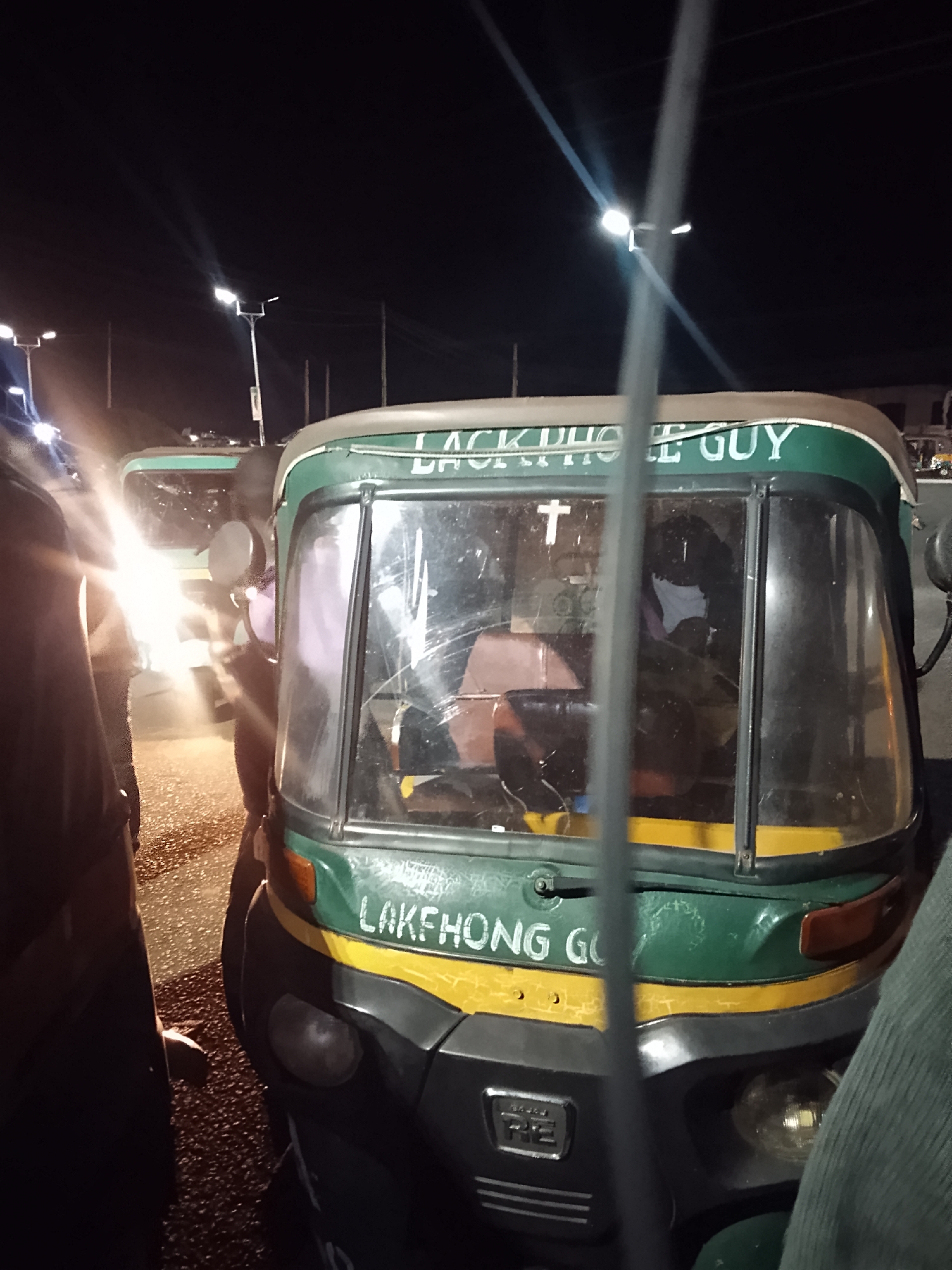 Here's a detailed description of the image:
**Scene:** The image appears to be taken at night in an urban environment. Streetlights are visible in the background.
**Subject:** The primary subject is a three-wheeled vehicle (likely an auto-rickshaw or "Keke Napep").
**Vehicle Details:**
* Color scheme: Green and yellow are prominent colors.
* Text: The vehicle has text on it. The top part reads "LACKPHOLE GUY". The second part reads "LAKEHONG GO".
* Condition: The vehicle appears to be in used condition, with some visible wear and tear. There is a cross attached to the windshield.
* Passengers: There appear to be passengers inside the vehicle.
**Environment:** The immediate environment looks like a street or roadside. There are other vehicles present in the background, along with other people.