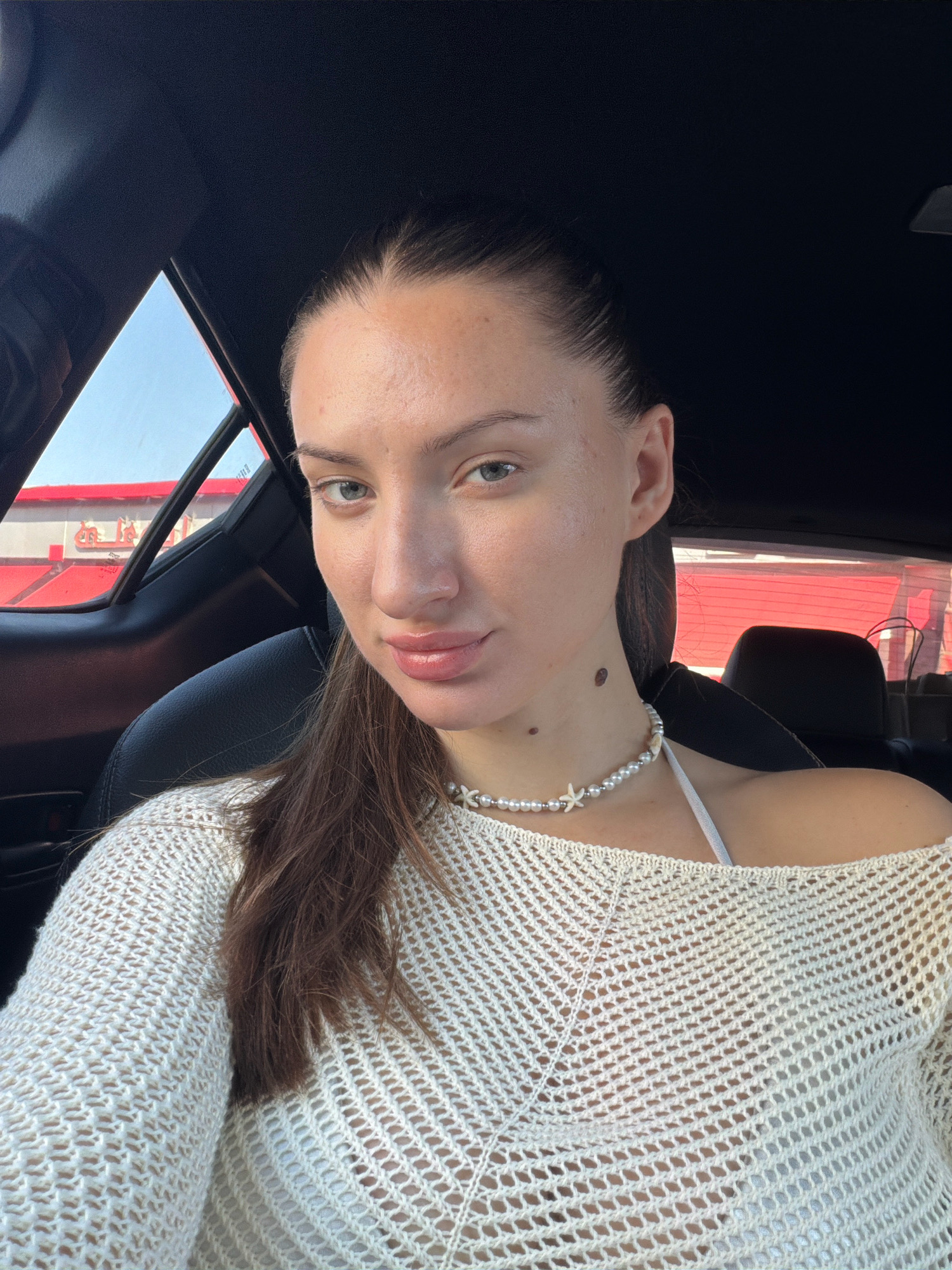 The image shows a young woman with long brown hair, fair skin, and light blue eyes, wearing a white, crocheted off-the-shoulder top and a pearl necklace with shell accents. She is sitting in a car, with the interior of the vehicle visible, including black leather seats and dark headliner. The background shows a building with a red facade and white lettering, suggesting a commercial establishment. The scene is likely set in a city environment, given the presence of buildings and a vehicle. There is no specific indication of Sharm El Sheikh, Egypt.