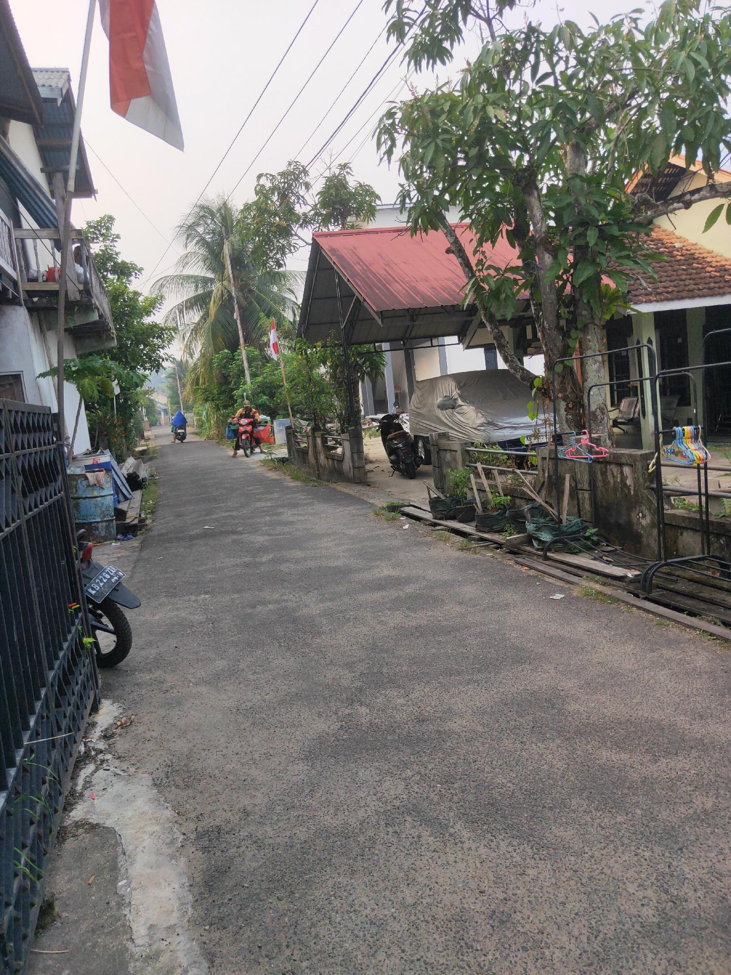 The image depicts a narrow asphalt road in a residential area, likely within the city of Sintang, Indonesia. The road is flanked by houses and vegetation. A partially visible motorcycle with license plate K 8228 XXJ is parked on the left, partially obscured by a black metal fence. Further down the road, two individuals are riding a red motorcycle, and another person is riding a blue motorcycle. A large tree with lush green leaves stands near the right side of the road, casting shadows. Several plants in pots are visible near the base of the tree. A car covered with a tarp is parked in front of a house on the right. In the background, a white building with a red-tiled roof is visible. An Indonesian flag, red and white, is flying on a pole on the left side of the road, near a balcony. The sky is overcast, suggesting a hazy or cloudy day. The scene appears to be a quiet, everyday moment in a community.