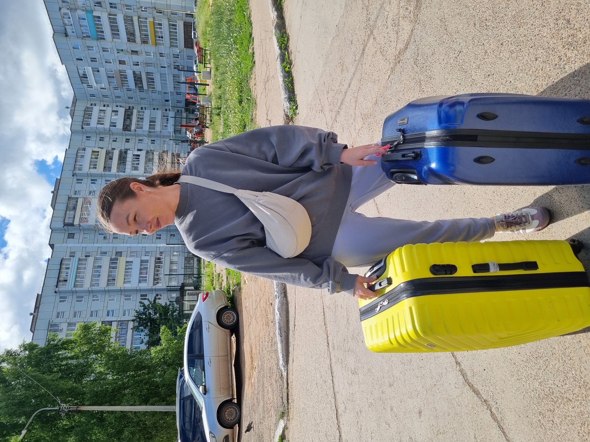 A woman stands on a paved area in front of a light-blue apartment building in Chaikovsky, Russia, holding two suitcases. The woman is facing to her right, looking away from the camera. She is wearing a grey sweatshirt, light grey sweatpants, and white and purple athletic shoes. A light-colored crossbody bag is slung across her chest. She is holding the handle of a bright yellow suitcase in her right hand and the handle of a dark blue suitcase in her left hand. The yellow suitcase is to her left, and the blue suitcase is to her right. The apartment building is several stories high with multiple windows and balconies. Behind the building, there is a grassy area with a playground visible. A silver car is parked to the left of the frame, and a tree with green foliage is to the far left. The sky is partly cloudy.