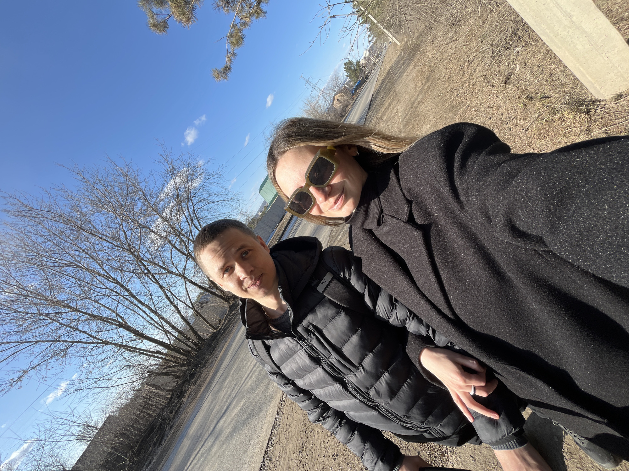 A selfie shows a man and a woman standing outdoors on a sunny day in what appears to be a roadside environment. The woman wears sunglasses and a black coat. The man wears a black puffer jacket. The road and surrounding landscape appear somewhat barren, suggesting early spring. Bare trees stand nearby, with a blue sky visible above. The image was taken in the city of Chaikovsky, Russia.