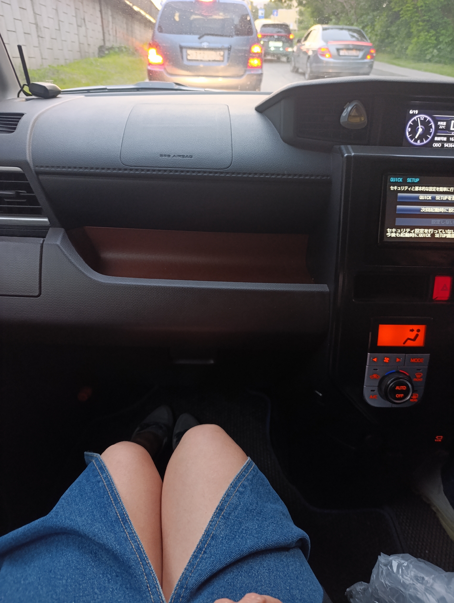 The image depicts the interior of a car, likely a passenger vehicle, from the perspective of someone sitting in the driver's or passenger's seat. The dashboard is visible, featuring air vents, a digital display showing the date "6/19" and other information, and climate control buttons. The foreground shows a person's legs, clad in a denim skirt, and feet wearing dark shoes. Outside the car, through the windshield, a line of traffic is visible on a road bordered by a concrete barrier and greenery, suggesting an urban or suburban setting. The specific location within Penza, Russia, cannot be determined from the visual information provided.