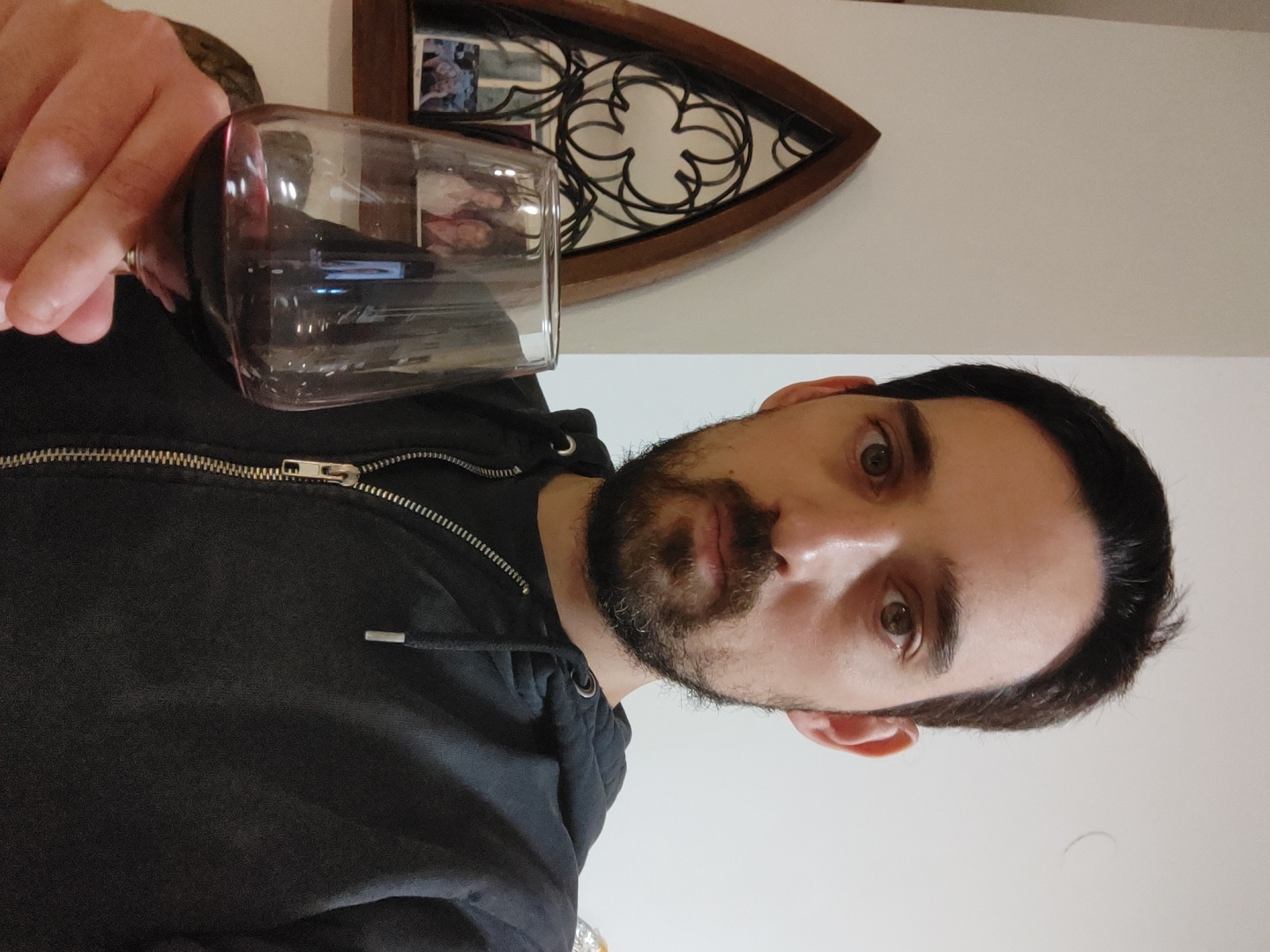 A bearded man in a black hooded jacket is holding a glass of red wine. Behind him is a decorative wall hanging of an arched window with a floral design. The background is a plain white wall. The location is unknown and cannot be determined from the image.