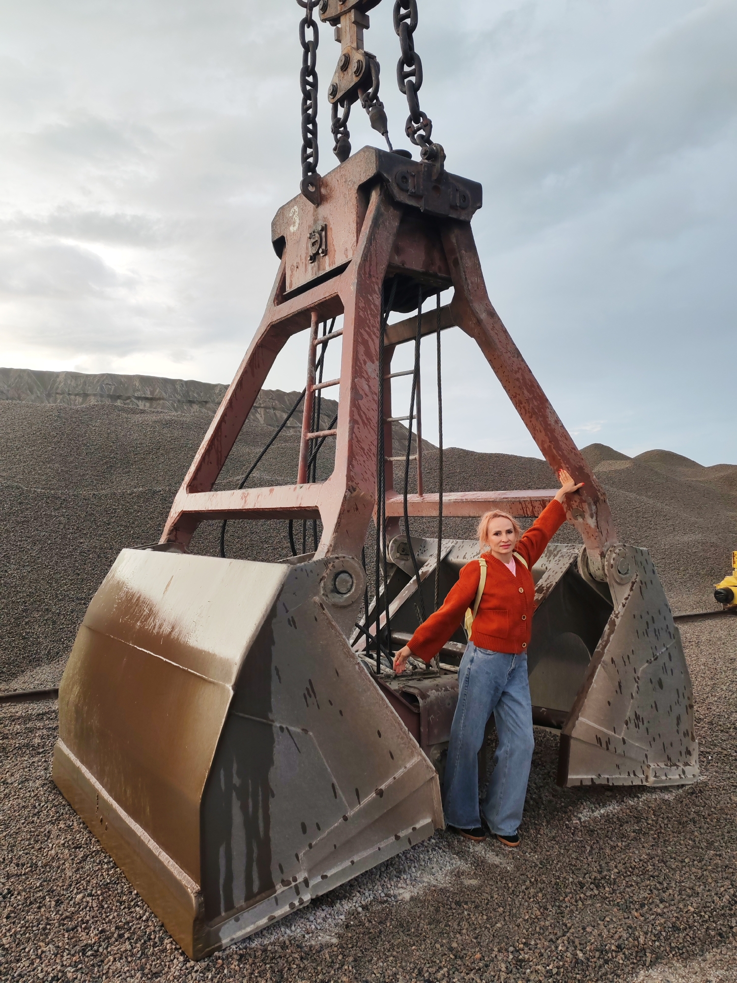A woman with pink hair, wearing an orange cardigan and blue jeans, stands in front of a large orange grab crane bucket on a gravel-covered surface. The crane bucket is a dominant feature, with heavy chains extending upwards. Behind the crane, piles of gravel stretch into the distance, forming undulating hills under an overcast sky. The location appears to be an industrial or quarry site, possibly in the city of Shalsky, Russia, indicated by the presence of heavy machinery and gravel stockpiles. The woman is positioned in a way that suggests she is posing for the photo, with her arms extended.