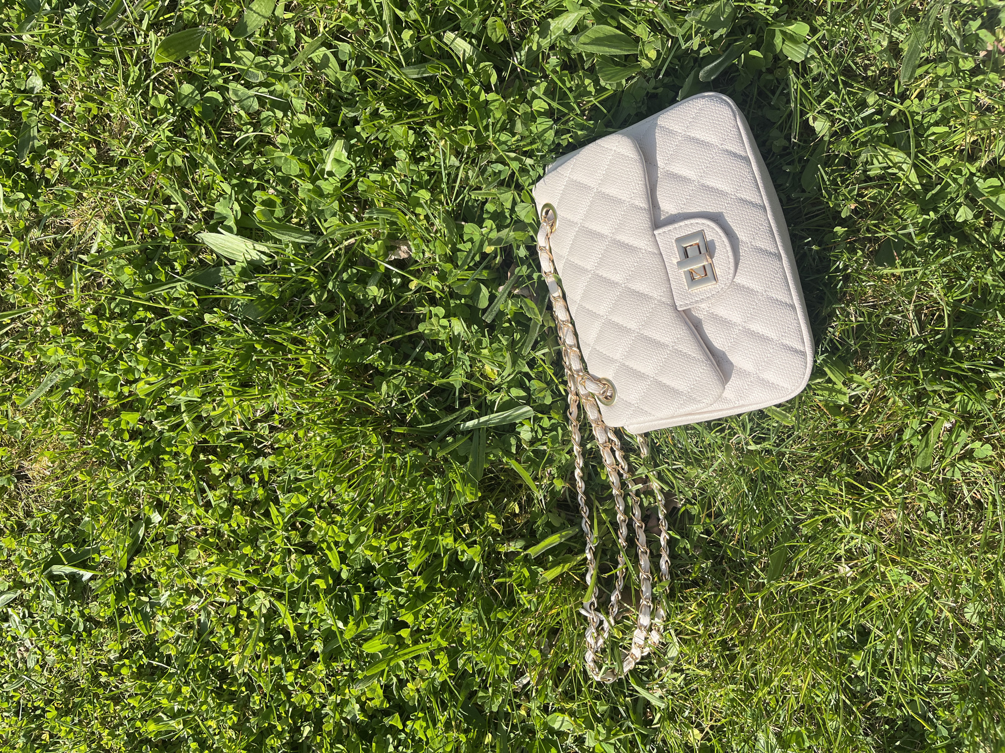 The image shows a white quilted purse lying on grass. The purse has a gold-colored chain strap. The location is unknown, but the request is for Varna, Bulgaria. The grass appears to be a mix of grass blades and clover. There are no people visible.