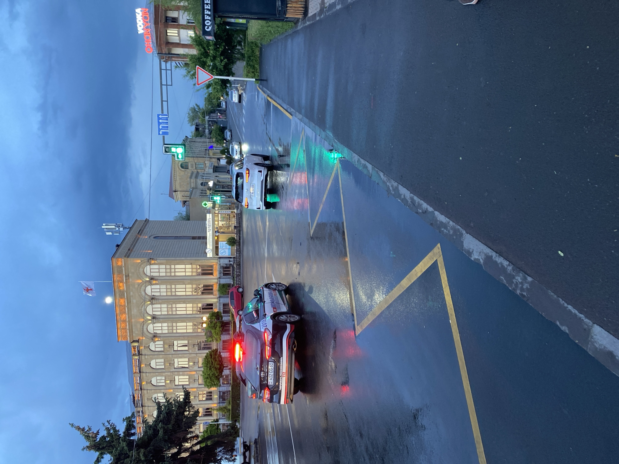 The image depicts a street scene in Gyumri, Armenia, at dusk. A police car with its emergency lights flashing is parked on the wet asphalt. The car, a black Toyota Camry, has license plate "412 CG 61". In the background, a large, illuminated building with classical architectural features stands prominently. Across the street, a "COFFEE" sign is visible on a kiosk. A traffic light shows a green signal, indicating that traffic is permitted to proceed. The sky is overcast with dark clouds, and the wet road surface reflects the lights of the vehicles and surrounding buildings. The overall impression is one of a city street after a rain shower, with a sense of ongoing activity due to the presence of the police car and moving traffic.