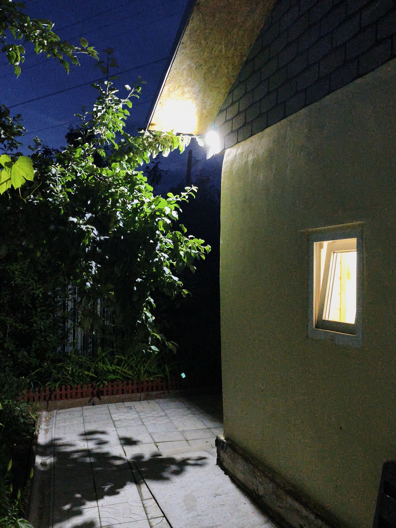 This image depicts an exterior view of a building at night. The building has a textured, light green stucco wall and a dark shingled roof. A small, lit window is visible on the wall, with yellow curtains partially obscuring the light. An outdoor light fixture illuminates the eaves of the roof and casts shadows on the surrounding area.
To the left of the building, there is a patio area paved with square tiles. Shadows of tree branches are cast across the patio, suggesting the presence of nearby foliage. A small, decorative wooden fence runs along the edge of the patio, separating it from a garden area with lush green plants. A tree with dense green leaves and some visible fruits is also present, with its branches extending towards the building and the sky. The sky is dark blue, indicative of nighttime, with a few faint horizontal lines that could be power lines.
The overall mood of the image is quiet and serene, with the warm light from the window and the outdoor fixture providing a sense of domesticity against the dark, natural backdrop. The time of day is clearly night. There is no discernible weather, but the darkness suggests no active precipitation. Location cues such as the architectural style of the building and the type of vegetation might suggest a temperate climate. No text is visible in the image.