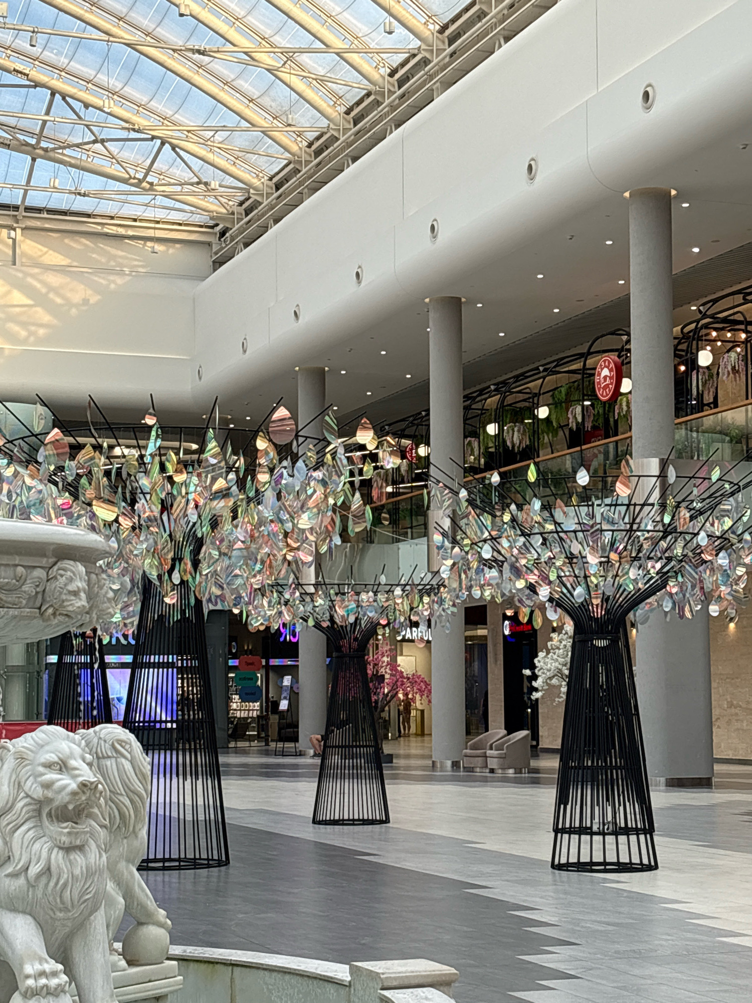 The image depicts the interior of a modern shopping mall, likely a public space within the city of Kyiv, Ukraine. The architecture features a high, translucent ceiling with a visible metal support structure, allowing natural light to filter in. Large, pale grey cylindrical columns punctuate the space. In the foreground, a white marble sculpture of two lions, one with its mouth open, sits in front of a fountain, suggesting a decorative element.
Dominating the mid-ground are three abstract tree-like sculptures constructed from black metal frames. These sculptures are adorned with numerous iridescent, leaf-shaped ornaments that reflect light and color, creating a vibrant, shimmering effect. Behind these sculptures, the mall environment is visible, with shops and storefronts lining the upper levels. Signs with text, some in Cyrillic script, are present. There are also some decorative elements like pink artificial trees and white artificial trees visible within the mall. The floor is a patterned grey and white tile.
The lighting in the scene suggests it is daytime, with a warm, natural light illuminating the space. There are no people prominently featured in the immediate foreground or mid-ground, though individuals may be present in the background within the shops or walking through the mall. The overall impression is one of a spacious, well-lit, and artistically decorated commercial environment.