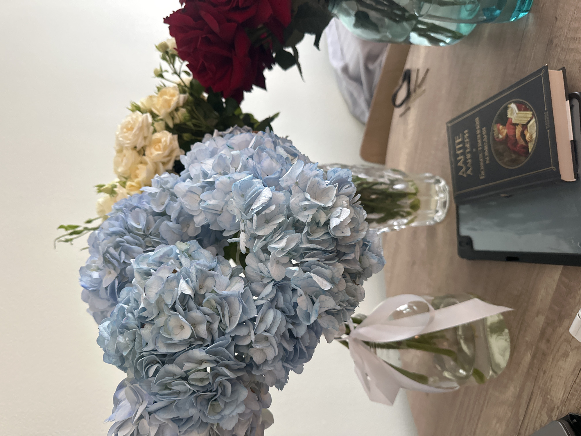 In the foreground, a dark-covered book lies open on a wooden surface, with a circular illustration depicting a figure reading. To the left of the book, a dark tablet is visible. Behind these items, a clear glass vase adorned with a light pink ribbon contains a bouquet of large, pale blue hydrangeas. Further back, a taller, clear, textured vase holds a bouquet of small, pale yellow roses. To the right, a vibrant red rose is partially visible, emerging from a turquoise glass vase. The background consists of a plain, light-colored wall. The scene appears to be an indoor setting, likely a home or a studio, given the presence of personal items and floral arrangements. There is no indication of the specific location in Moscow, Russia, or any activity taking place.
