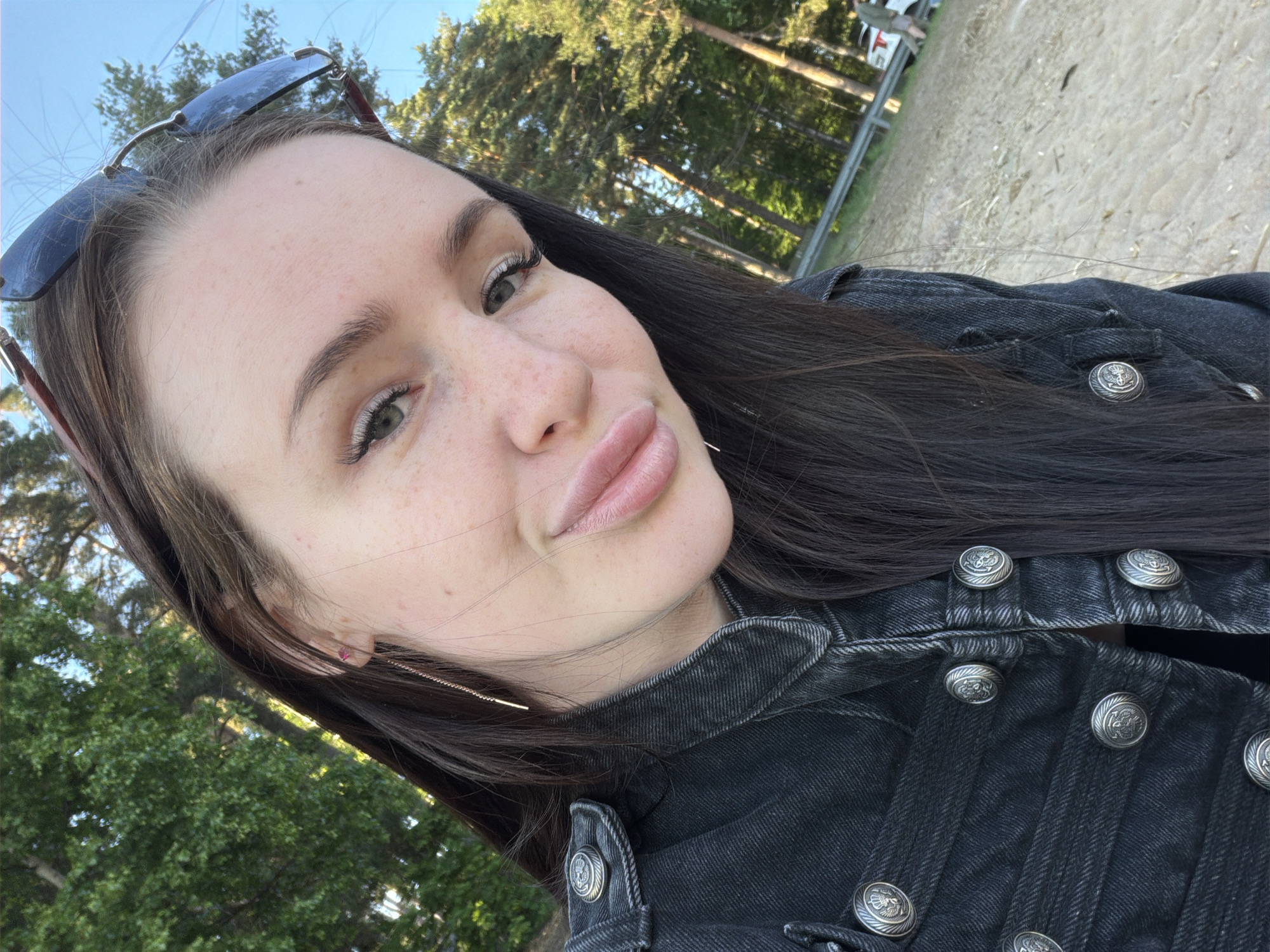 An outdoor close-up portrait of a young woman is presented in Repino, Russia. The woman, who appears to be in her twenties or early thirties, has long, dark brown hair, light-colored eyes, and visible freckles across her nose and cheeks. She wears small, pinkish-red stud earrings in both ears, and long, gold-toned threader earrings. Dark-lensed sunglasses with brown frames are positioned on top of her head. She is wearing a dark grey or black denim jacket featuring a military-style design with multiple large, circular, silver-toned buttons embossed with an ornate crest or eagle. The background reveals a clear blue sky above tall evergreen trees with dense green foliage. Below the trees, a sandy or dirt area is visible, bordered by a silver metal guardrail. In the distant right background, a white vehicle and a person in light clothing are partially visible. The woman is looking directly at the camera with a subtle smile.