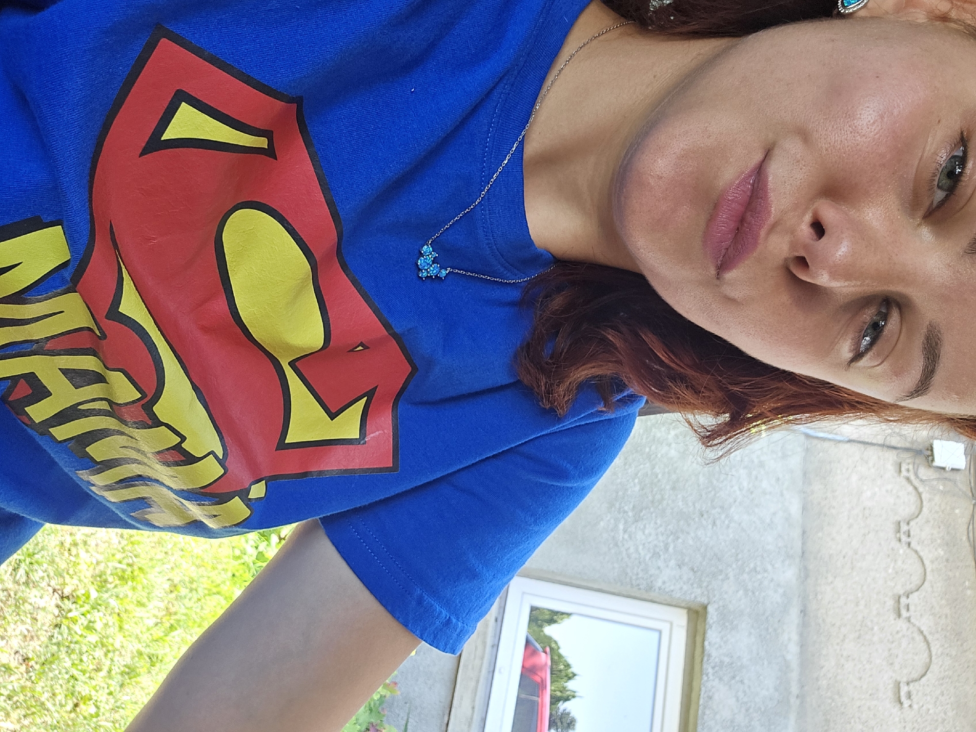 A woman with reddish-brown hair and green eyes is pictured from the chest up, wearing a bright blue t-shirt with a Superman logo. The logo is predominantly red and yellow, with "MAMA" written in yellow letters below it. She is wearing a delicate silver necklace with a cluster of small blue stones. The background shows a textured, light-colored wall with decorative arches and a window reflecting trees and a red car. The scene appears to be outdoors, with green grass and foliage visible in the lower right corner, suggesting a sunny day. The location is identified as Sofia, Bulgaria.