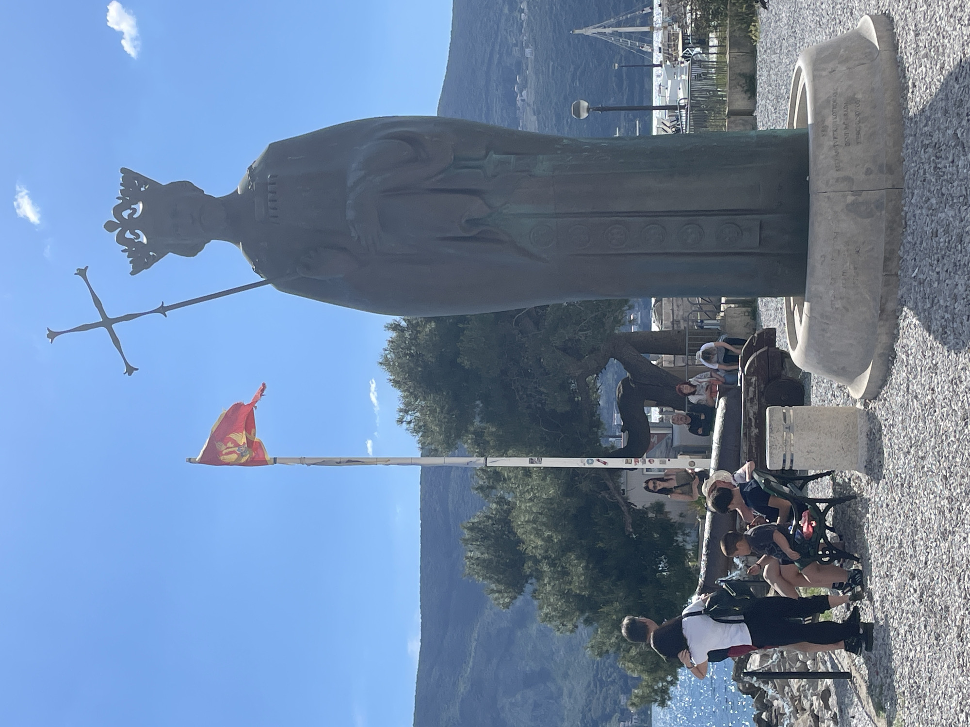 The image, oriented correctly, displays a large, weathered bronze statue of a crowned figure in an outdoor setting in Herceg Novi, Montenegro. The majestic statue, likely depicting a historical monarch or saint, wears an ornate robe and holds a staff topped with a crucifix in its right hand. It stands on a circular, light-colored stone base.
In the mid-ground, several individuals, including adults and children, are present. Some are seated on green park benches, while others stand around. Behind them, a large, dark-colored historical cannon or similar armament is visible. A tall flagpole prominently displays a red and yellow flag adorned with an emblem, possibly a local or historical Montenegrin symbol.
The background features a sprawling, tree-covered mountain range under a brilliant, clear blue sky dotted with a few small white clouds. To the right, elements of a harbor, including boats and distant structures, can be seen, hinting at the coastal location. The ground in the immediate foreground is covered with light-colored gravel.
The scene takes place during bright daylight, suggesting a sunny midday or early afternoon. People appear relaxed, enjoying the outdoor environment.
Text is inscribed in Cyrillic on the statue's base. Clearly visible phrases include "СВЕТИ СТЕФАН" (Saint Stephen), "КРАЉ СРБА" (King of Serbs), and "ПРВОВЕНЧАНИ" (The First-Crowned), indicating the statue is dedicated to Stefan the First-Crowned. Additional Cyrillic text is present on the left side of the base but is not legible.