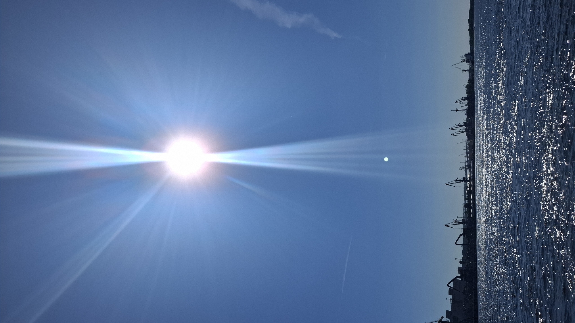 The image shows a bright sun with prominent rays against a clear blue sky. Below the sky, a dark silhouette of a cityscape is visible, featuring numerous cranes, suggestive of a port or industrial area. The foreground is occupied by dark blue water with a shimmering surface due to sunlight reflection. A faint contrail is visible in the sky to the right of the sun, and another thin line, possibly a contrail or a cloud streak, is present on the left. The overall scene suggests a sunny day at a port. This scene is consistent with the appearance of the port facilities located on Vasilievsky Island in Saint Petersburg, Russia, with the sun being a dominant element in the sky.