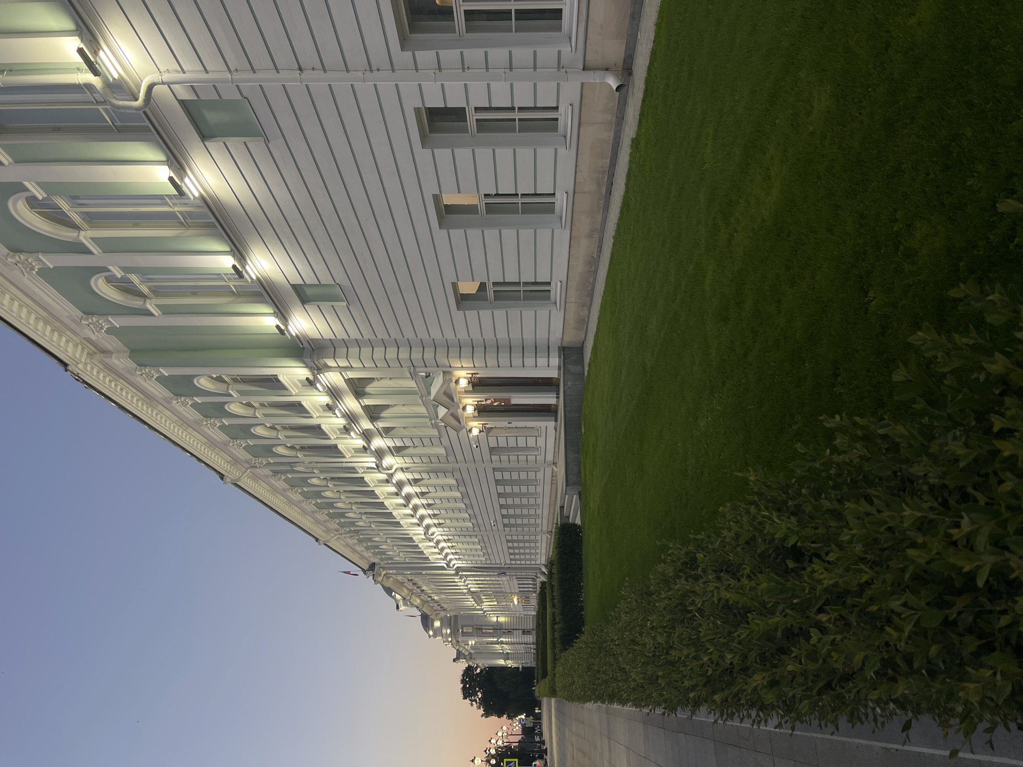 The image shows a long, neoclassical building illuminated by exterior lights at dusk. The building is painted white and light green, with a series of arched windows and decorative architectural elements. A manicured green lawn stretches in front of the building, bordered by a paved walkway on the left. In the distance, streetlights and trees are visible along the walkway. The sky is a pale blue, indicating the end of daylight. This building is the Tver State Technical University's main building, located in Tver, Russia. There are no people or animals visible in the image.