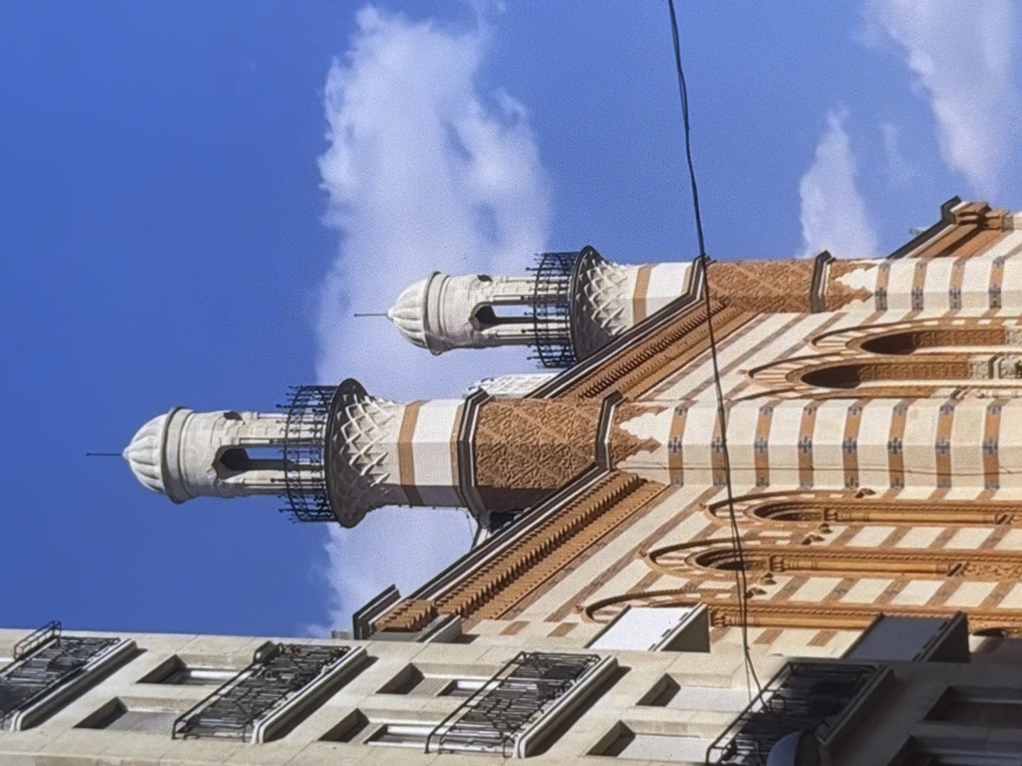 The media file is a photograph taken from a low angle, looking up at a building with two prominent minaret-like structures. The sky is a bright blue with scattered white clouds. The building itself features decorative architectural elements, including striped patterns in beige and orange, intricate lattice work around the upper sections of the minarets, and arched window-like openings. Below the main building, another structure with several windows and metal railings is visible. A thin black wire runs diagonally across the upper right portion of the image. The lighting suggests it is daytime, and the clear sky indicates fair weather. No people or specific activities are depicted. The overall impression is of an ornate building against a clear sky, possibly in a city.