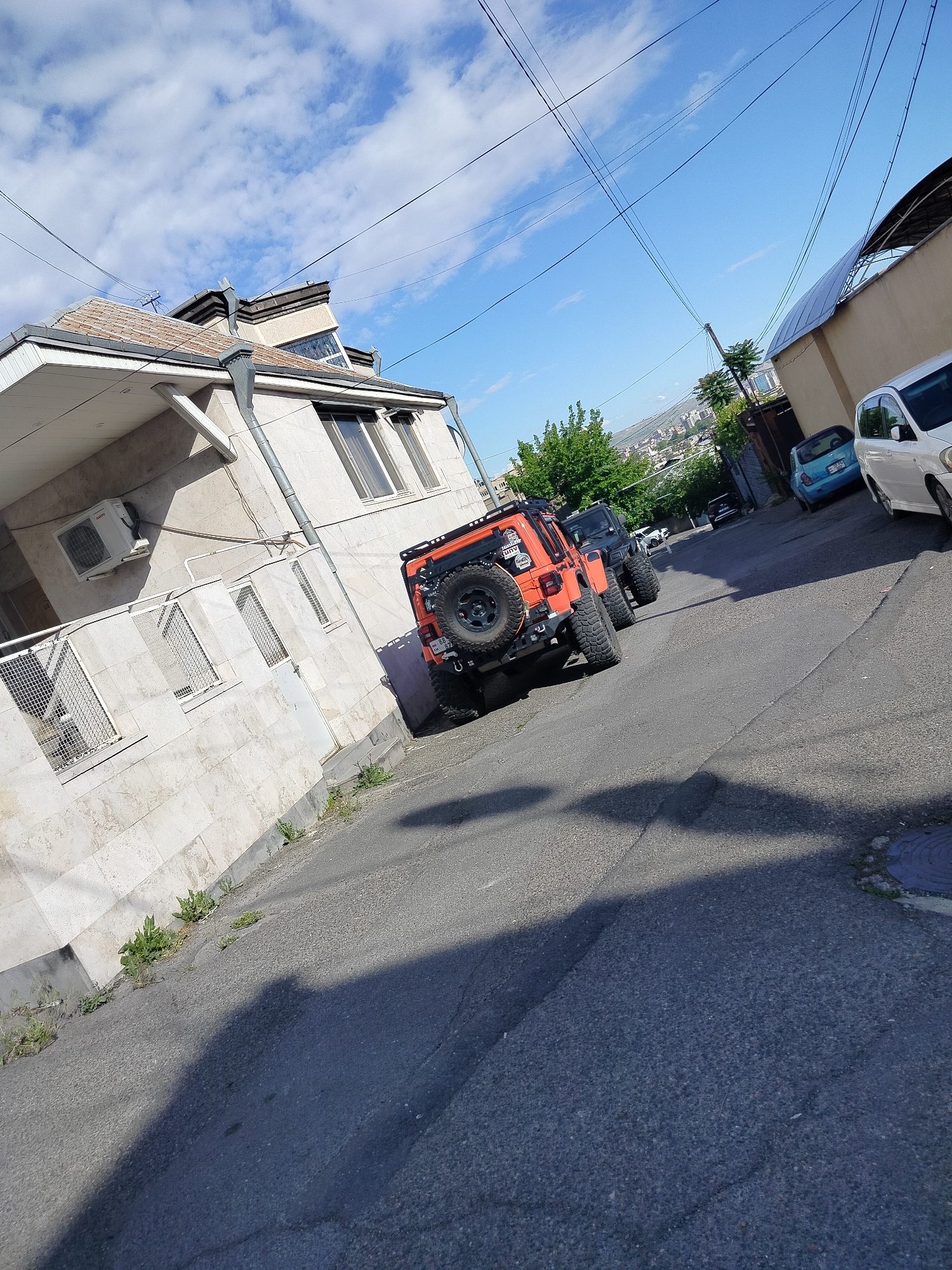 The image shows a street scene in Yerevan, Armenia, with an orange Jeep parked along the side of the road. Adjacent to the Jeep is a light-colored stone building with an air conditioning unit visible. Further up the inclined street, other vehicles are parked, including a black Jeep and two cars on the right. The sky is partly cloudy, and electrical wires are strung overhead. The street is paved and appears to be residential, with buildings lining both sides. In the background, there are distant hills and possibly the city skyline. The scene appears to be captured during the daytime, with sunlight casting shadows on the street. A manhole cover is seen in the bottom right corner of the image.
