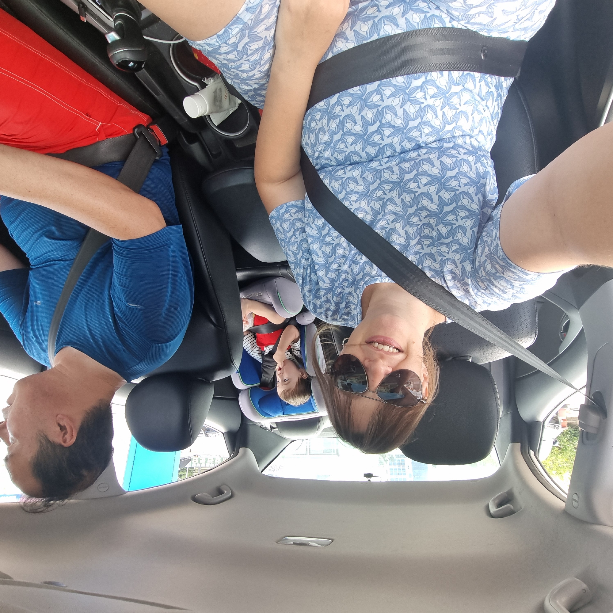 In Gangneung-si, South Korea, a family is captured in a selfie taken from the back seat of a car, likely during a road trip. The image shows a woman in the foreground wearing sunglasses and a blue and white patterned dress. She is smiling and appears to be the one taking the photo. Behind her, a toddler sits in a car seat, wearing a striped shirt and red shorts, looking towards the side. To the right of the woman, a man wearing a blue t-shirt and red shorts is visible, looking forward, presumably driving. The car interior is discernible, with black leather seats and seatbelts prominently featured, highlighting safety precautions. The background outside the car windows suggests an urban environment with buildings and trees, consistent with being in a city. The overall impression is a candid moment of a family enjoying a journey together.