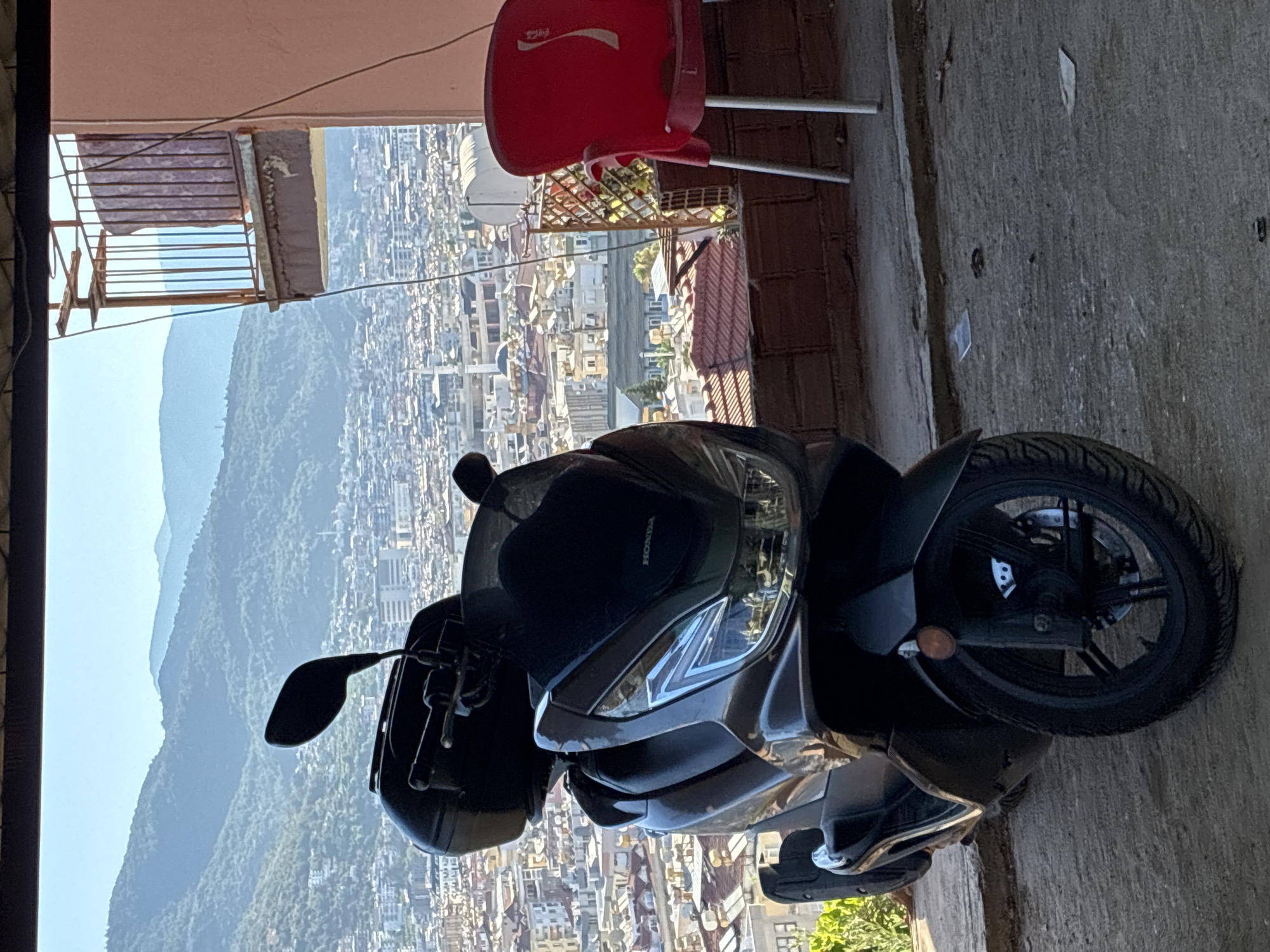 The image depicts a black Honda PCX 125 scooter parked on a concrete surface with a scenic view of Alanya, Turkey in the background. The scooter is positioned in the foreground, dominating the left side of the frame. It is a modern scooter with a black seat, black storage box mounted on the rear, and prominent front headlights. The scooter's handlebars and mirrors are visible.
Behind the scooter, the cityscape of Alanya unfolds. Numerous buildings with red-tiled roofs are densely packed, extending towards a backdrop of lush green mountains. The hazy silhouette of distant mountains suggests a clear day with good visibility. A partial view of a building with a red Coca-Cola branded chair and a balcony with a metal railing is visible on the right side of the image. The overall lighting indicates it is daytime, likely late morning or early afternoon, given the bright sunlight and shadows. The scene appears to be from a vantage point, possibly a terrace or balcony, overlooking the city. There are no people visible in the image. The ground beneath the scooter is concrete and appears somewhat worn, with some debris scattered on it.