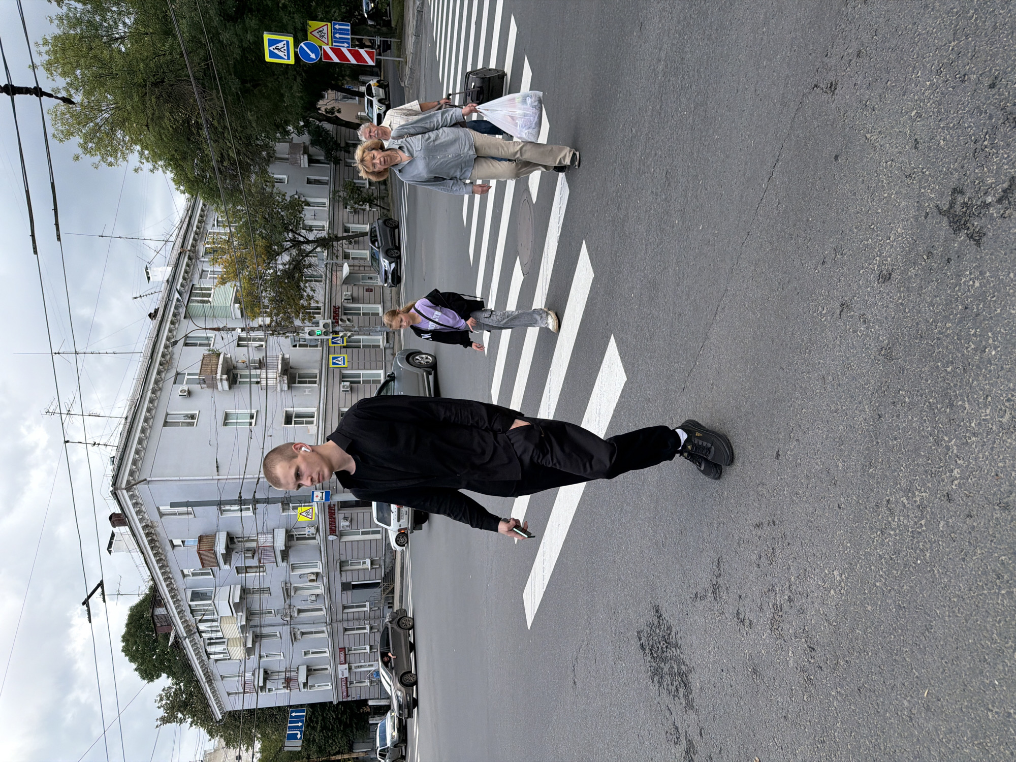 A young man with short hair and white earbuds is crossing a pedestrian crossing in Nizhny Novgorod, Russia, on an overcast day. He is wearing a black long-sleeved shirt and black pants, with black athletic shoes. He is holding a smartphone in his right hand. Behind him, a young girl in a purple top and black jacket is also crossing the street, followed by an older woman in a light gray shirt and beige pants, carrying a black duffel bag and a plastic bag filled with groceries. An older man is walking behind the woman. In the background, a light blue apartment building with multiple balconies and trees line the street. Traffic lights and street signs are visible. Cars are driving on the street.