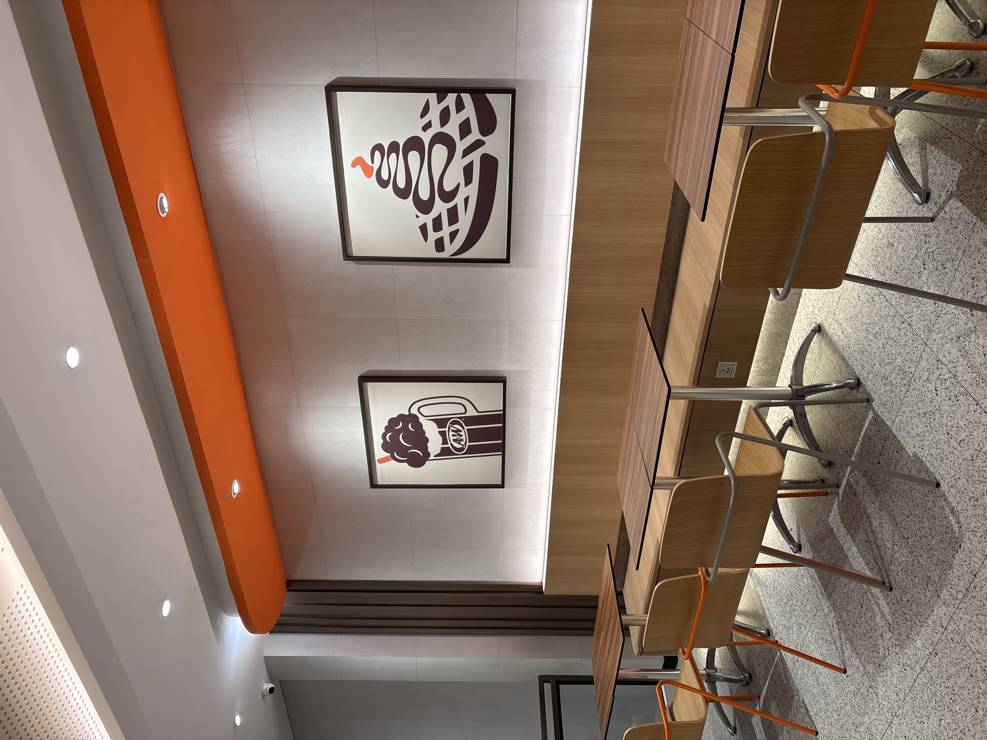 A food establishment interior features a modern seating area with two framed artworks depicting a root beer float and a waffle with ice cream. The walls are tiled in a light grey, with a light-colored wooden bench seat running along the wall. Two-seater tables with dark wood tops and metal bases are arranged in front of the bench. Several wooden chairs with metal legs and orange accents are placed around the tables. The floor is a speckled light grey tile. The ceiling has recessed lighting and a prominent orange soffit. The establishment appears to be a fast-food restaurant. The image location is identified as Sukabakti, Indonesia. No people are present in the image.