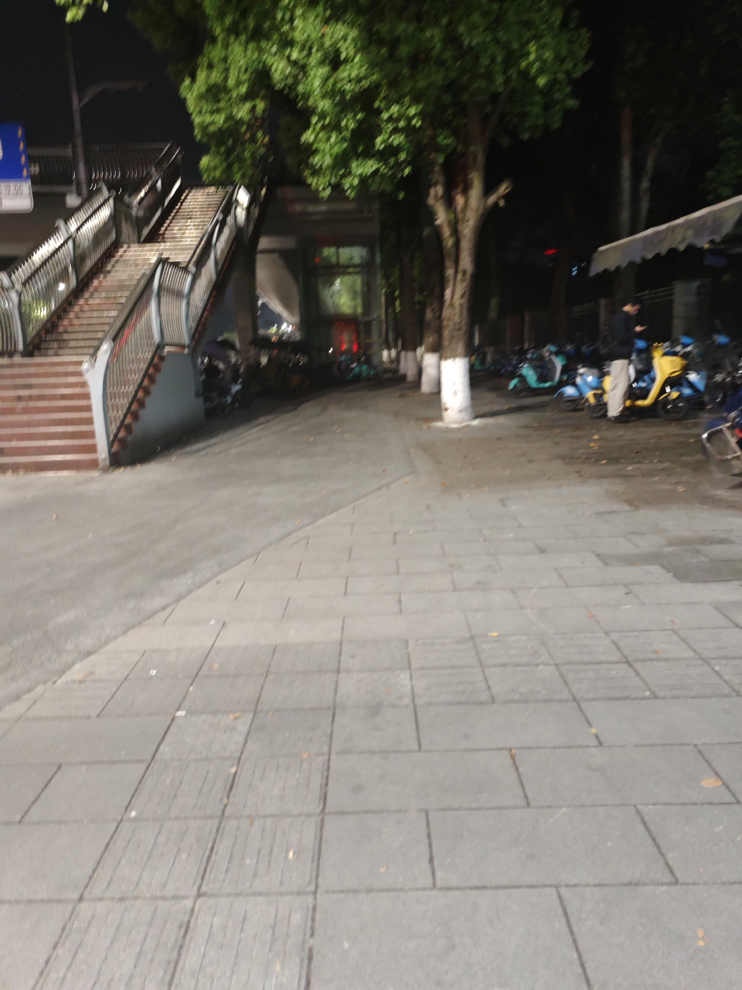 The image shows a street scene at night in Chongqing High-tech Zone, China. A pedestrian overpass is visible on the left side of the image. Several electric scooters are parked under an awning on the right, where a person stands looking at a phone. Trees line the street, and their trunks are painted white near the base. The ground is paved with square, gray stones.