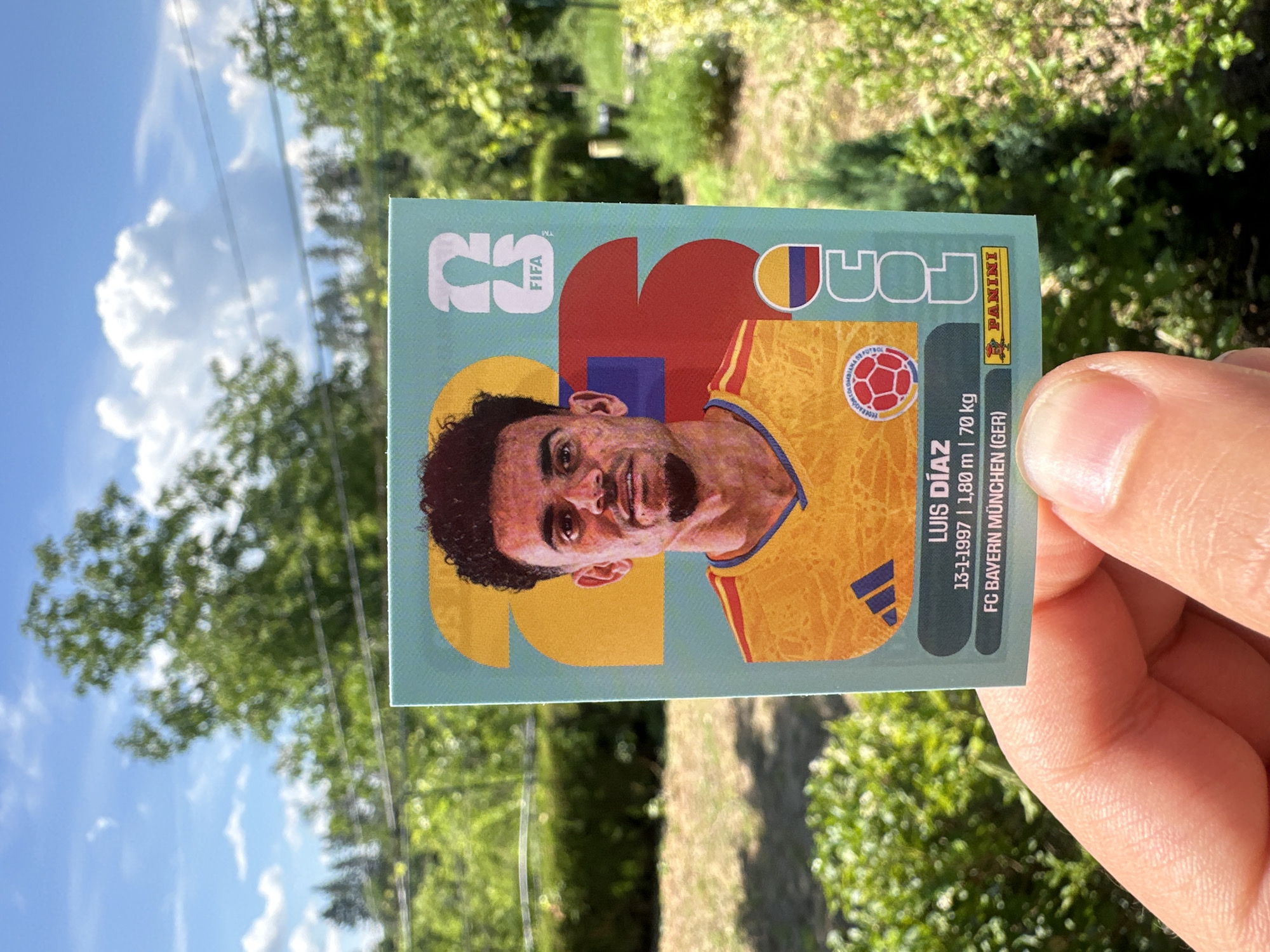 This is a close-up, outdoor, eye-level shot of a person's hand holding a trading card. The card features a portrait of a man with dark curly hair and a beard, wearing a yellow jersey with blue and red accents. He has a slight smile and his head is tilted. The card has text that includes "FIFA", "PANINI", and "LUIS DÍAZ". Below his name are his birthdate, height, and weight, followed by his club, "FC BAYERN MÜNCHEN (GER)". The background of the card is a light teal color with abstract shapes in red, yellow, and blue.
The background of the image is blurred but shows a sunny outdoor scene with green trees and a bright blue sky with white clouds. The lighting suggests it is daytime. The setting appears to be a natural environment, possibly a park or garden, given the lush greenery. The composition focuses on the trading card and the hand holding it, with the background out of focus.