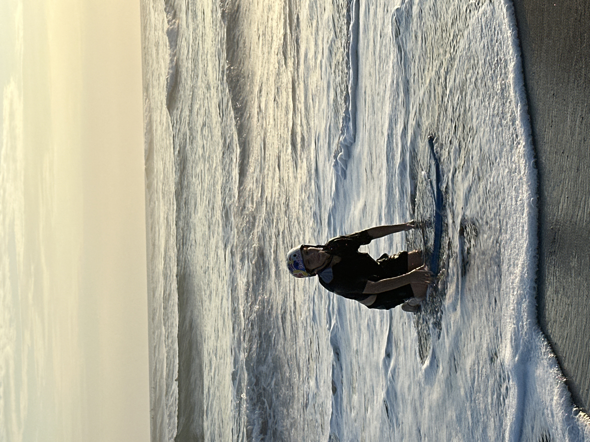A person wearing a helmet and dark clothing kneels on a boogie board in shallow ocean water. The person is facing the viewer, with waves breaking around them. The background consists of the ocean and a hazy sky. The location is the beach in North Kuta, Indonesia.