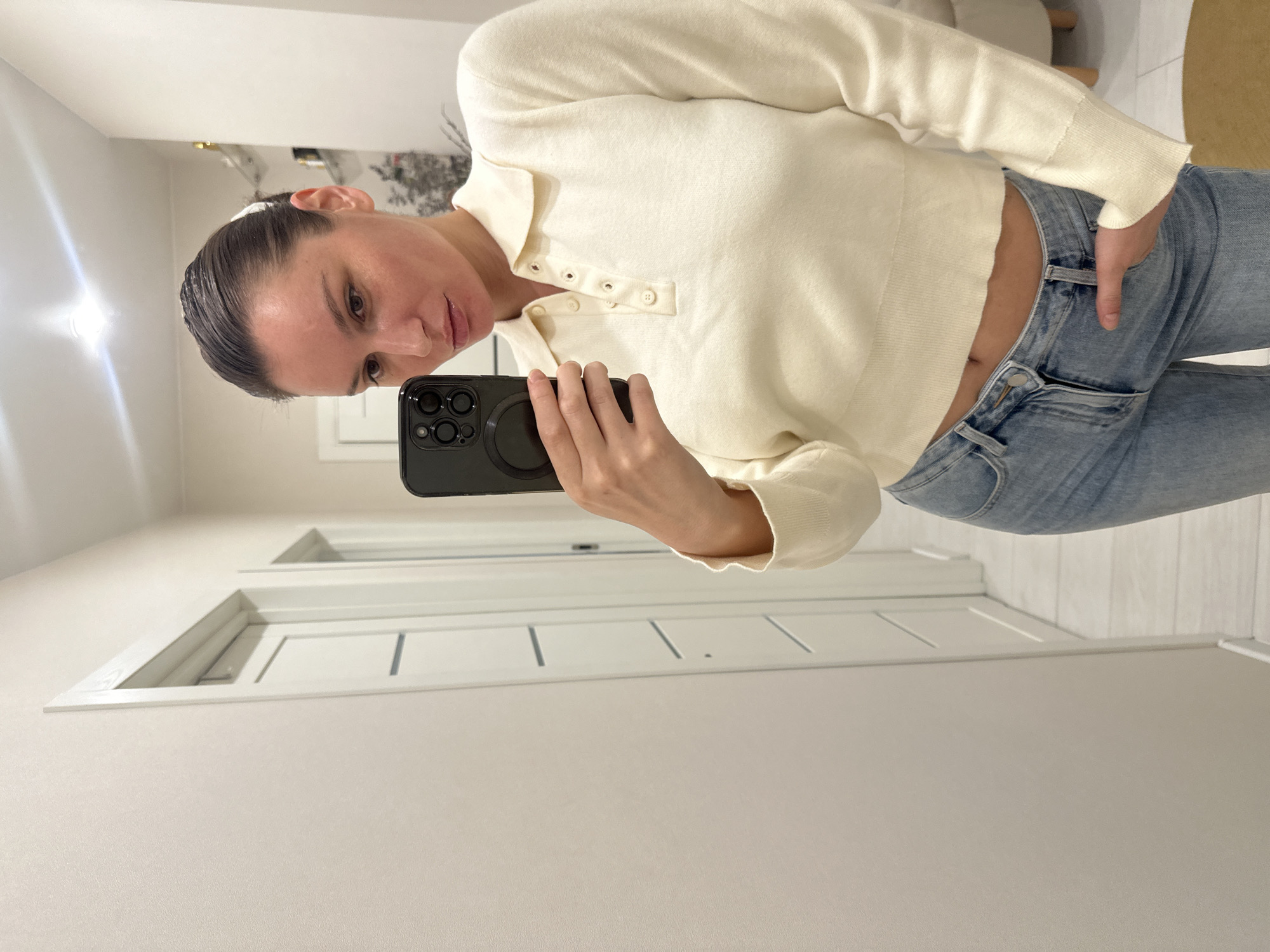 A woman is captured in a mirror selfie, her reflection appearing in an indoor setting. She has dark hair, slicked back from her face, which is tilted slightly and shows a neutral to contemplative expression. She is wearing a light cream or off-white long-sleeved knit sweater with a collared neckline and a three-button placket, revealing a small portion of her midriff. Her left hand is casually tucked into the front pocket of light blue denim jeans. She holds a black smartphone with multiple camera lenses and a circular magnetic accessory on its back in her right hand, directing it towards the mirror to take the picture.
The background, visible in the reflection, depicts a brightly lit interior space, likely a hallway or corridor. The walls are a light, neutral color. Several white doors with simple paneling details are visible along the wall behind her. A bright light source is reflected in the upper left corner of the image, suggesting artificial indoor lighting. The floor also appears light-colored. The clean, modern aesthetic of the room suggests a contemporary home or apartment in Moscow, Russia. No visible text is present in the image.