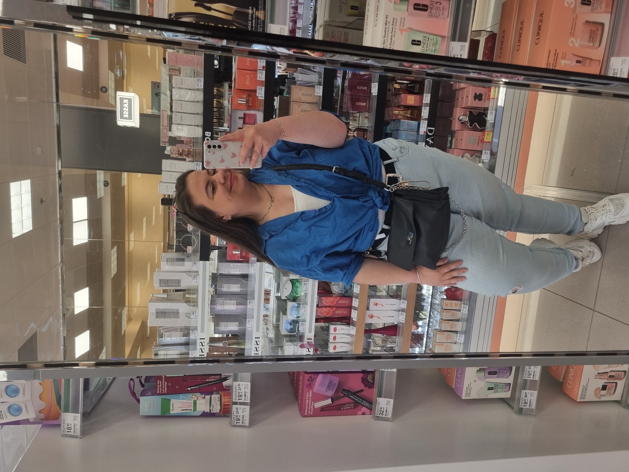 The image shows a woman taking a selfie in a mirror, likely inside a store. In the reflection, rows of cosmetics and skincare products are visible, including brands like Clinique and Dr. Jart+. Price tags indicate the location is in Europe, specifically Saarbrücken, Germany. The woman is wearing a blue button-up shirt, light-wash jeans, and white sneakers, and has a black crossbody bag.