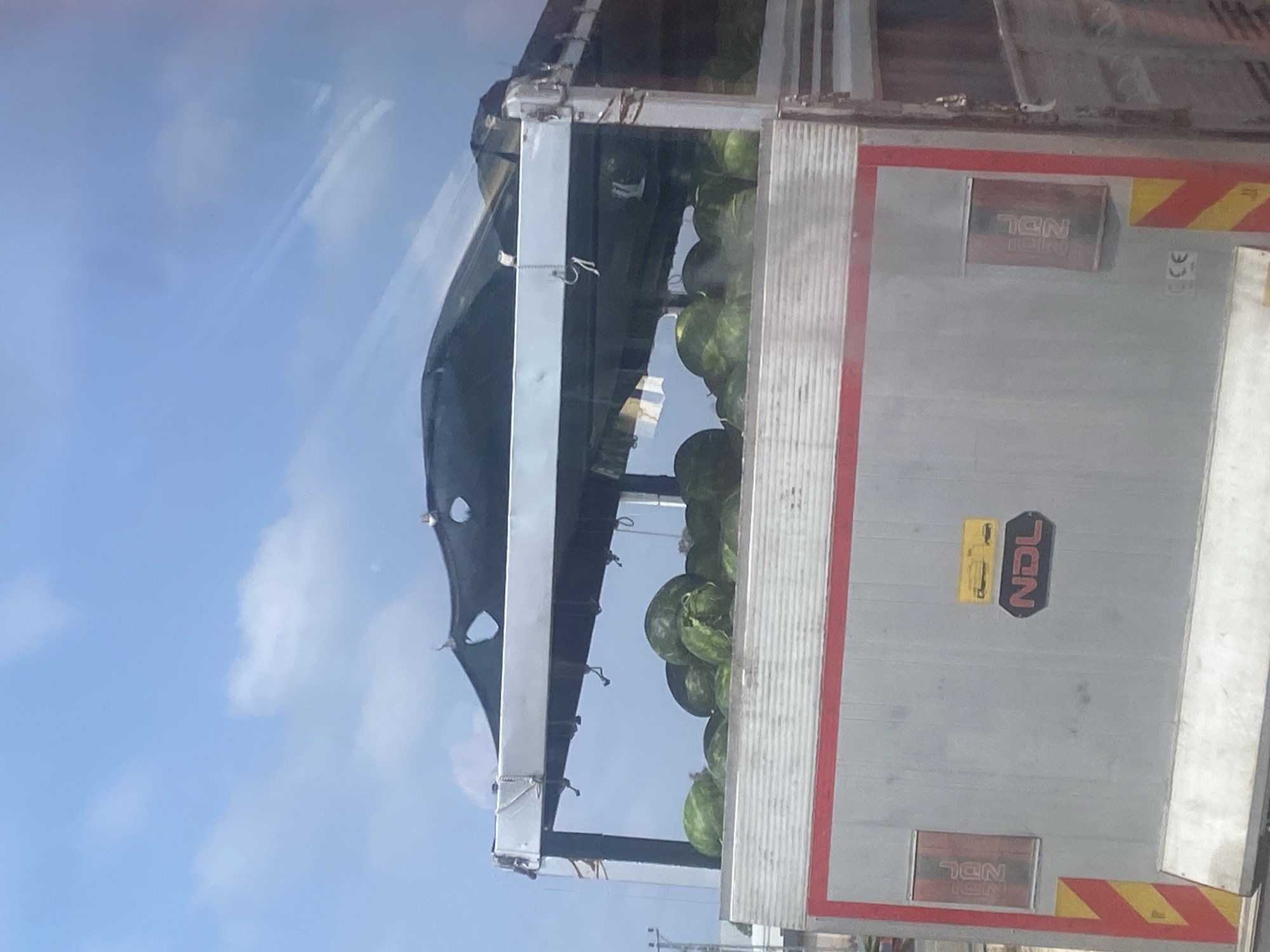 A commercial truck is filled with watermelons. The truck is silver with a red stripe and has the logo "NDL" on its side. The truck appears to be parked outdoors, possibly on a road or at a loading dock, under a blue sky with scattered clouds. The watermelons are green and fill the truck bed. The scene is likely in Serik, Turkey, a region known for watermelon cultivation.