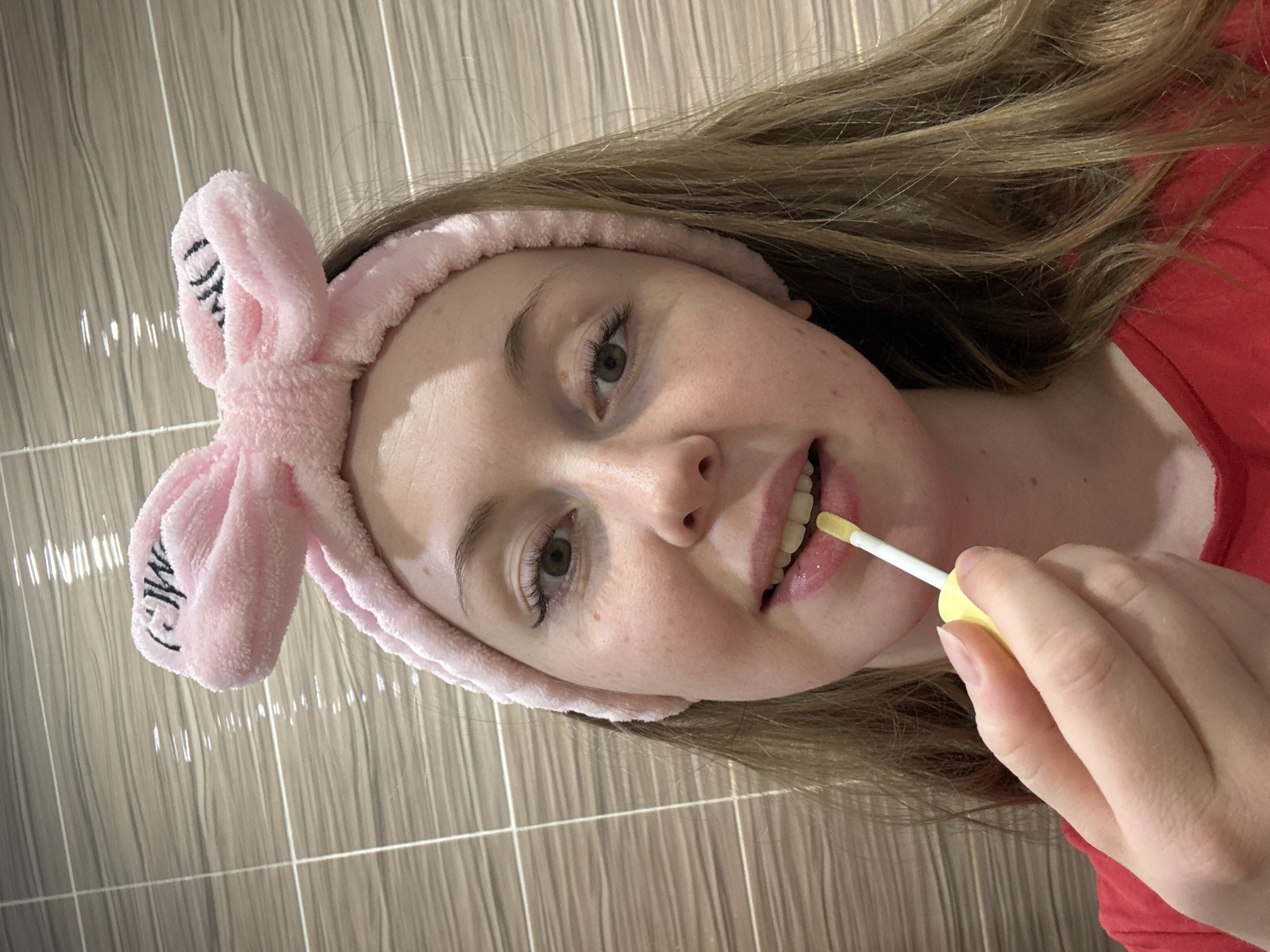 The image shows a young woman applying lip gloss. She is wearing a pink headband with bunny ears. Her hair is blonde, and she is wearing a red shirt. The background appears to be a bathroom with tiled walls. The location is Unknown, Unknown.