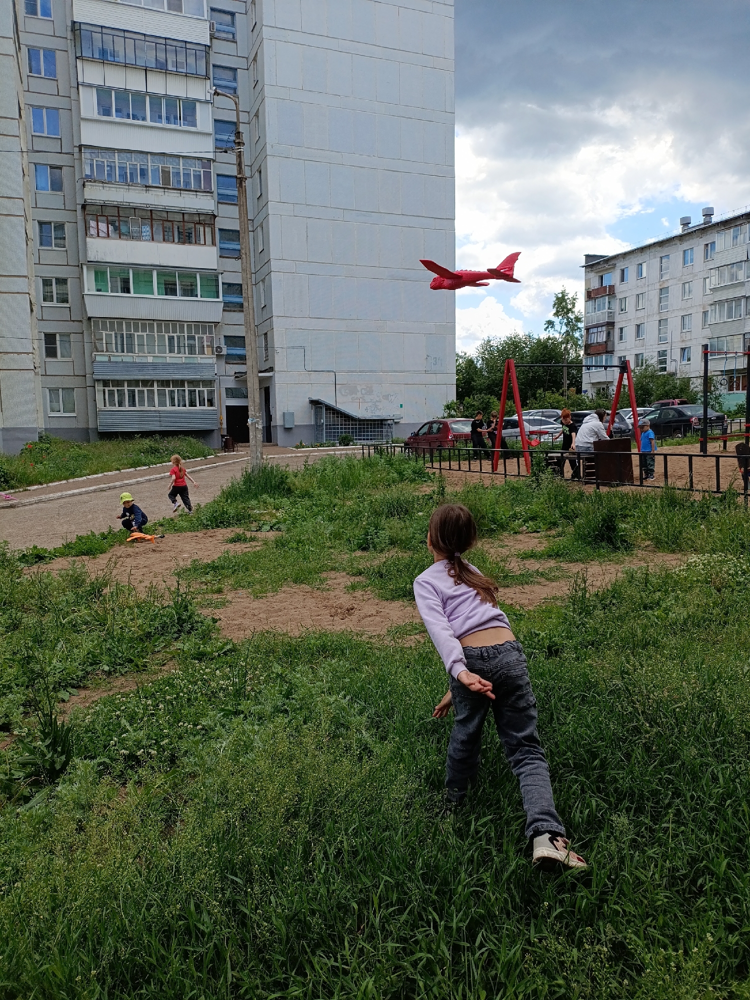 This detailed description of the image does not include any specific location information for Chaikovsky, Russia, as none is discernible within the visual content.
The image captures an outdoor daytime scene within a residential area featuring multiple apartment buildings, a grassy unkempt area, and several individuals, predominantly children, engaged in various activities.
In the foreground, a young girl with brown hair tied in a ponytail, wearing a light purple long-sleeved top and dark jeans, stands with her back partially to the viewer, facing slightly to the left. Her arms are bent behind her back, and she appears to be observing the scene ahead. The immediate ground beneath her is covered in dense green grass with some small white flowering plants.
The midground features a dirt patch surrounded by grass, transitioning into a paved asphalt path with a light-colored curb. On this path, a child with reddish hair, wearing a red t-shirt and light pants, is in a running stance. Another child, wearing a green cap and dark clothing, is crouched next to an orange scooter on the edge of the path.
In the background, on the left, a large multi-story grey concrete panel apartment building rises prominently. It features numerous windows and various styles of enclosed balconies. A utility pole with a light fixture is attached to the right side of this building. Another similar apartment building is visible on the far right, also with multiple windows and balconies, some appearing older. Between these buildings, against a partially cloudy sky with patches of blue, a bright red toy airplane with distinct wings and tail is suspended mid-air.
Further into the midground on the right, beyond the paved path, is a playground area enclosed by a low black metal fence. Several individuals, appearing to be young adults or older children, are gathered around playground structures, including red metal A-frame supports for swings and a dark, possibly wooden, climbing structure. One person in a white hoodie and another in a black t-shirt are visible within this group. Several parked cars (red, grey, black) are visible behind the playground fence.
The overall environment suggests a casual, possibly summer or late spring day, with ample daylight under a sky that is largely overcast but not uniformly so. The ground is a mix of natural elements, including overgrown grass and exposed dirt/sand, typical of an urban or suburban recreational space.