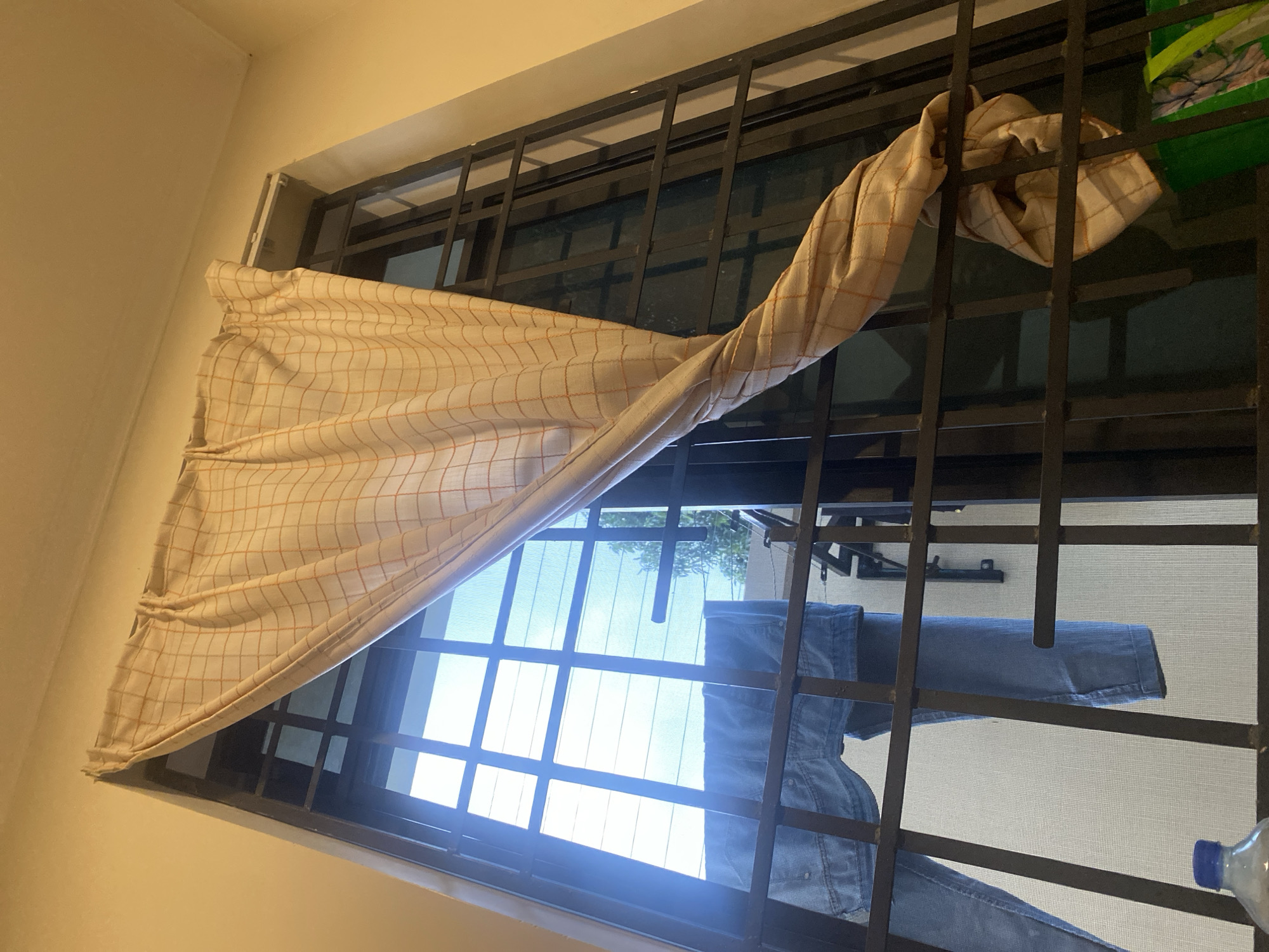 The image captures an indoor perspective looking out through a window with dark metal security bars. A light-colored fabric, possibly a curtain or cloth with a beige base and a fine orange-brown checkered pattern, is draped over the top section of the security grille, creating soft folds. The fabric extends from the upper left to the upper right of the frame.
Bright daylight streams through the window, indicating it is daytime, likely a sunny morning or afternoon. Beyond the security bars, a bright blue sky is visible, along with glimpses of green foliage from trees or bushes in the distance. Blue denim-like clothing, possibly jeans or a shirt, is hanging to dry on a line or rack outside the window, positioned behind the security bars. In the upper right corner, a portion of a green plastic object, possibly a bag with a floral or abstract pattern, is visible. The lower right corner reveals the top of a clear plastic water bottle. The interior walls around the window are a plain, light-colored (cream or pale yellow) surface. The presence of security bars and clothes drying outside a window are common sights in residential areas, aligning with the location context of Dar es-Salaam, Tanzania. There is no visible text in the image.