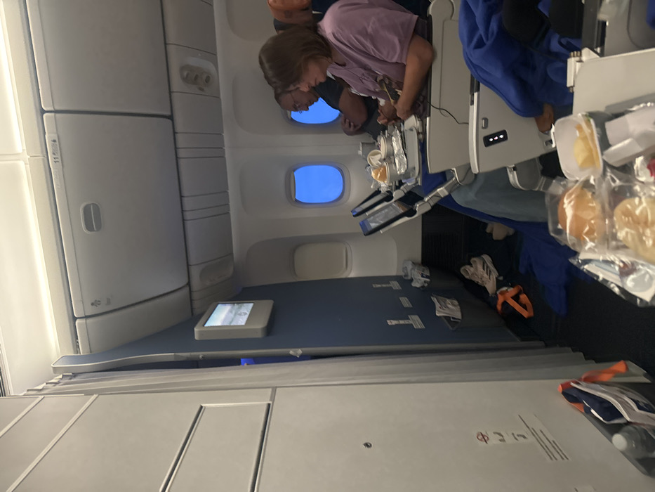 This image captures a sideways view inside an airplane cabin, appearing to be taken from a passenger's seat looking forward and slightly across the aisle. If oriented correctly, the image would show the cabin from left to right.
In the middle ground of the image, two passengers are visible seated next to each other by a window. The person closer to the camera (on the right side if the image were upright) is a woman with light brown hair, wearing a light purple short-sleeved top. She is leaning forward, looking down at something on her tray table, where an in-flight meal or snack is laid out. Her hands are engaged with the items on the table. Behind her and slightly to the left is another passenger, partially obscured, also looking down. Both appear to be engaged in an activity at their seats.
The seating arrangement suggests a modern aircraft, possibly a premium economy or standard long-haul configuration, with individual screens attached to the seatbacks or armrests. One such screen is visible on the blue surface in the lower section of the image (which would be the floor or side panel if upright), displaying what looks like an entertainment menu. Another screen is visible directly in front of the two seated passengers.
Through the two circular airplane windows, a clear, bright blue sky is visible, indicating that it is daytime and the plane is at cruising altitude.
Various objects are scattered around the cabin. On the tray table of the woman in purple, there's a small white container with a yellow substance (possibly chips or a side dish) and some bread rolls or pastries in clear plastic packaging. On the armrest or table closer to the camera (on the right), more packaged bread rolls are visible, along with what appears to be a plastic water bottle. Below the seats, on the floor, there are personal belongings, including a pair of light-colored athletic shoes (possibly white with pink accents) and a strap from an orange bag. Papers and wrappers are also scattered on the floor and seat areas.
Overhead compartments run along the top left side of the image (which would be above the aisle if upright). On one of these compartments, a small white sticker with black text, including "11" and other symbols, is faintly visible. Further down, near the bottom right, another white sticker with black text shows "8 L" or "8 A" with additional symbols, likely indicating a seat row and position or emergency instructions.
The overall scene depicts passengers relaxing during a daytime flight, likely enjoying a meal or using in-flight entertainment.