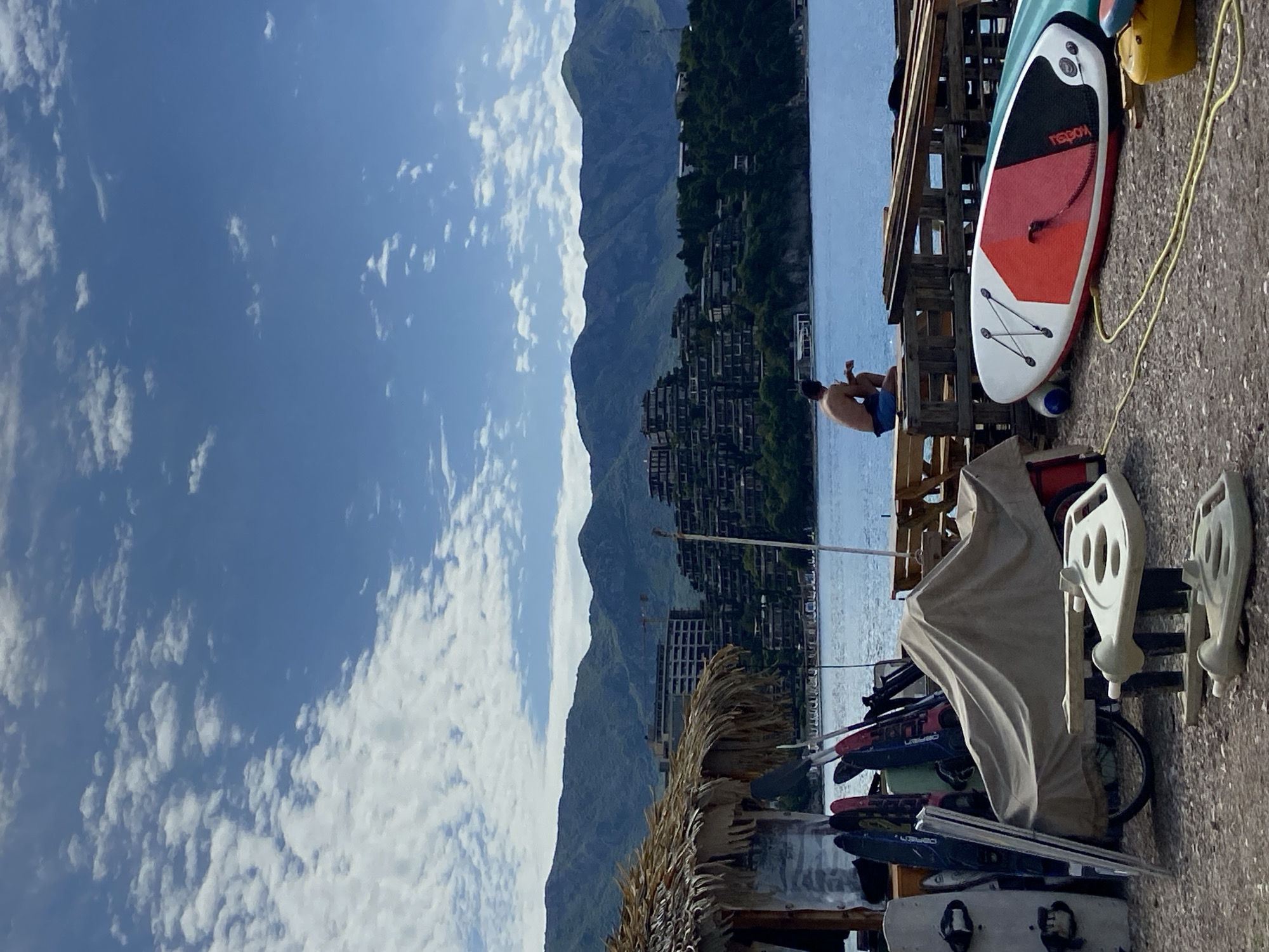 The image shows a view in Budva, Montenegro. In the background, there's a mountainous terrain partially obscured by clouds. Along the water’s edge, there are buildings and vegetation. A person is sitting at the edge of a wooden structure, looking out at the water. The foreground shows watersports equipment, including paddleboards and kiteboards, along with some other covered items. The sky is blue with scattered clouds. The ground appears to be concrete or pavement.