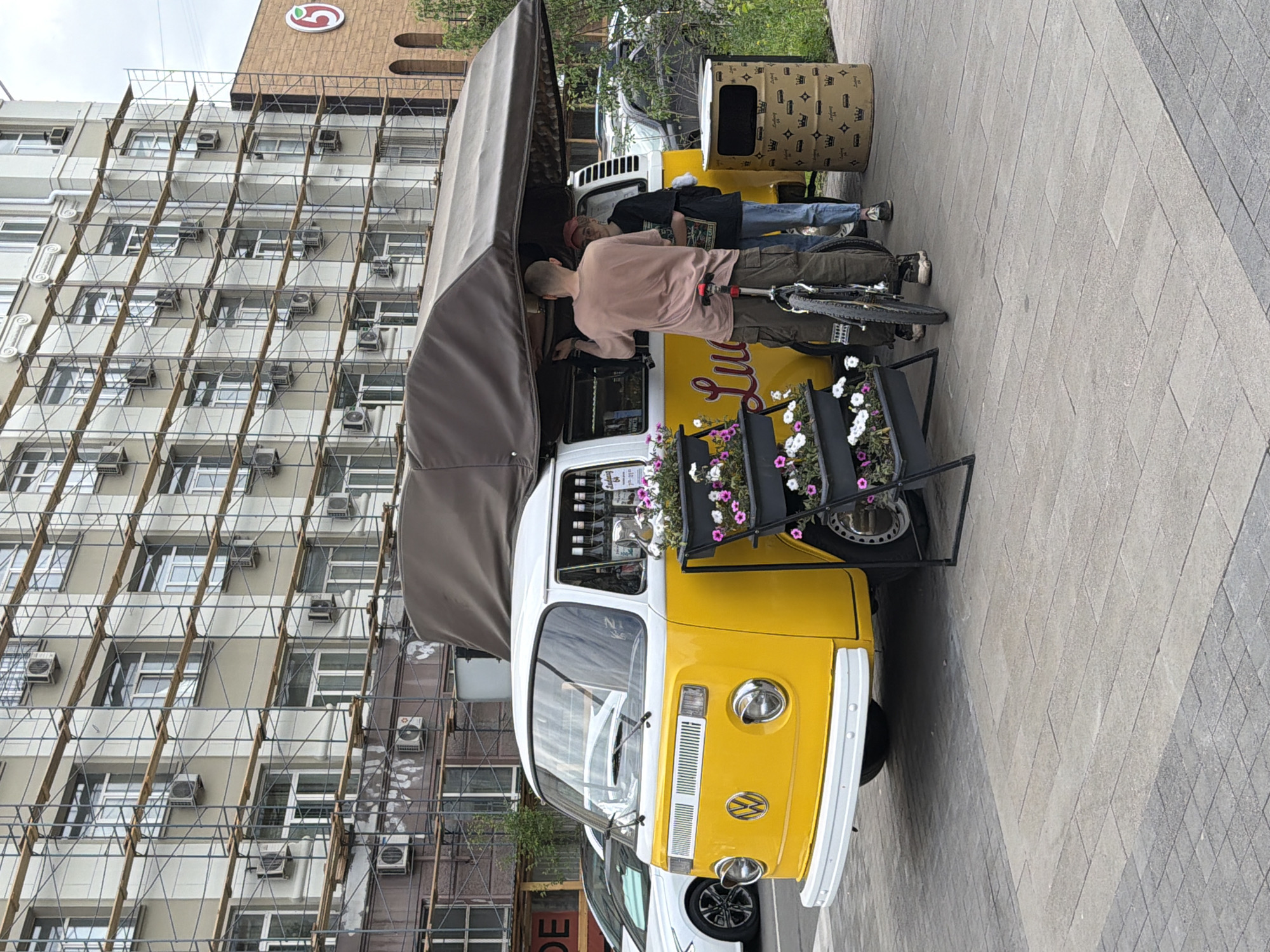 A yellow and white Volkswagen Type 2 van, modified into a food truck, is parked on a paved area in front of a multi-story building undergoing renovation, indicated by scaffolding covering its facade. The building is on the right side of the frame. A white car is parked behind the food truck on the left.
Two individuals, a man and a woman, are standing next to the food truck. The man, wearing a light brown t-shirt and olive green cargo pants, is leaning against a bicycle parked between him and the food truck. The woman, wearing a black t-shirt and blue jeans, is standing to the man's right. Both appear to be interacting with the food truck.
The food truck has a large brown canopy extended over its serving window. Several bottles are visible inside the serving area. A planter with pink and white flowers is placed in front of the food truck. A cylindrical trash receptacle, decorated with a crown pattern, stands to the right of the food truck.
The ground is paved with rectangular stone tiles. The background shows a modern building with many windows and air conditioning units, with scaffolding erected on its exterior. A red logo is visible on the building on the right side of the image. This scene is likely located in the city of Krasnoyarsk, Russia, given the context of the surrounding architecture and style.