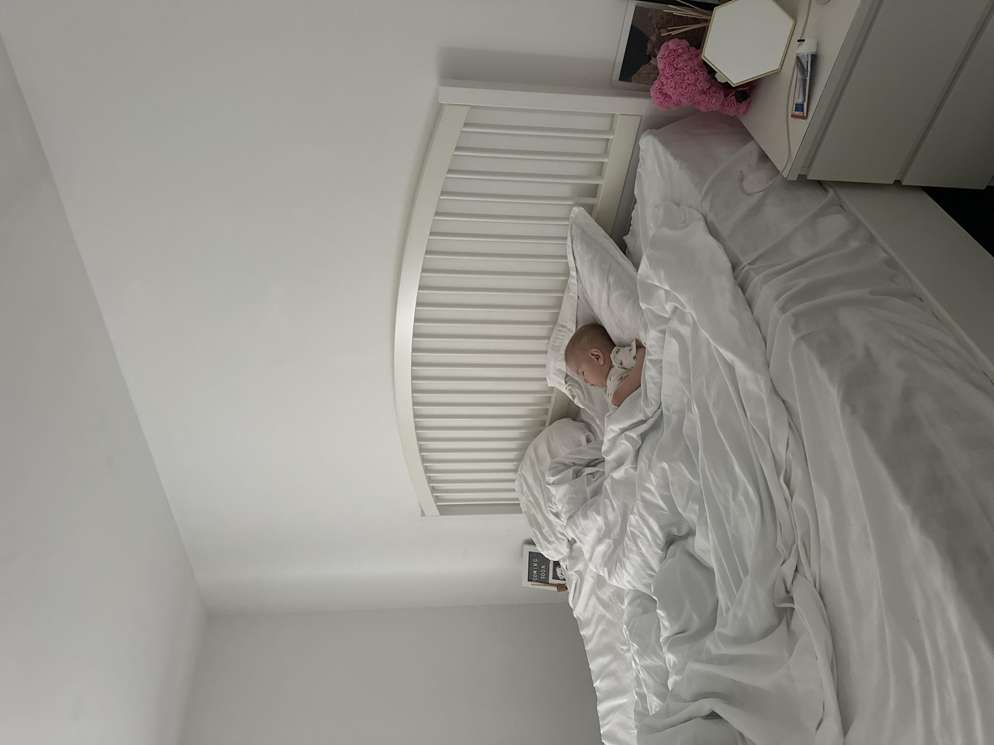 A baby sleeps soundly in a large bed with white linens. The bed is positioned against a white headboard, which has vertical slats. The room appears to be a bedroom with white walls and a white nightstand next to the bed. On the nightstand, there is a framed picture, a pink rose bear decoration, a hexagonal mirror, and a tube of toothpaste. To the left of the bed, a small framed sign that reads "COMING SOON" is visible on the floor. The overall impression is one of a peaceful, domestic environment. This scene is located in Kutenholz, Germany.