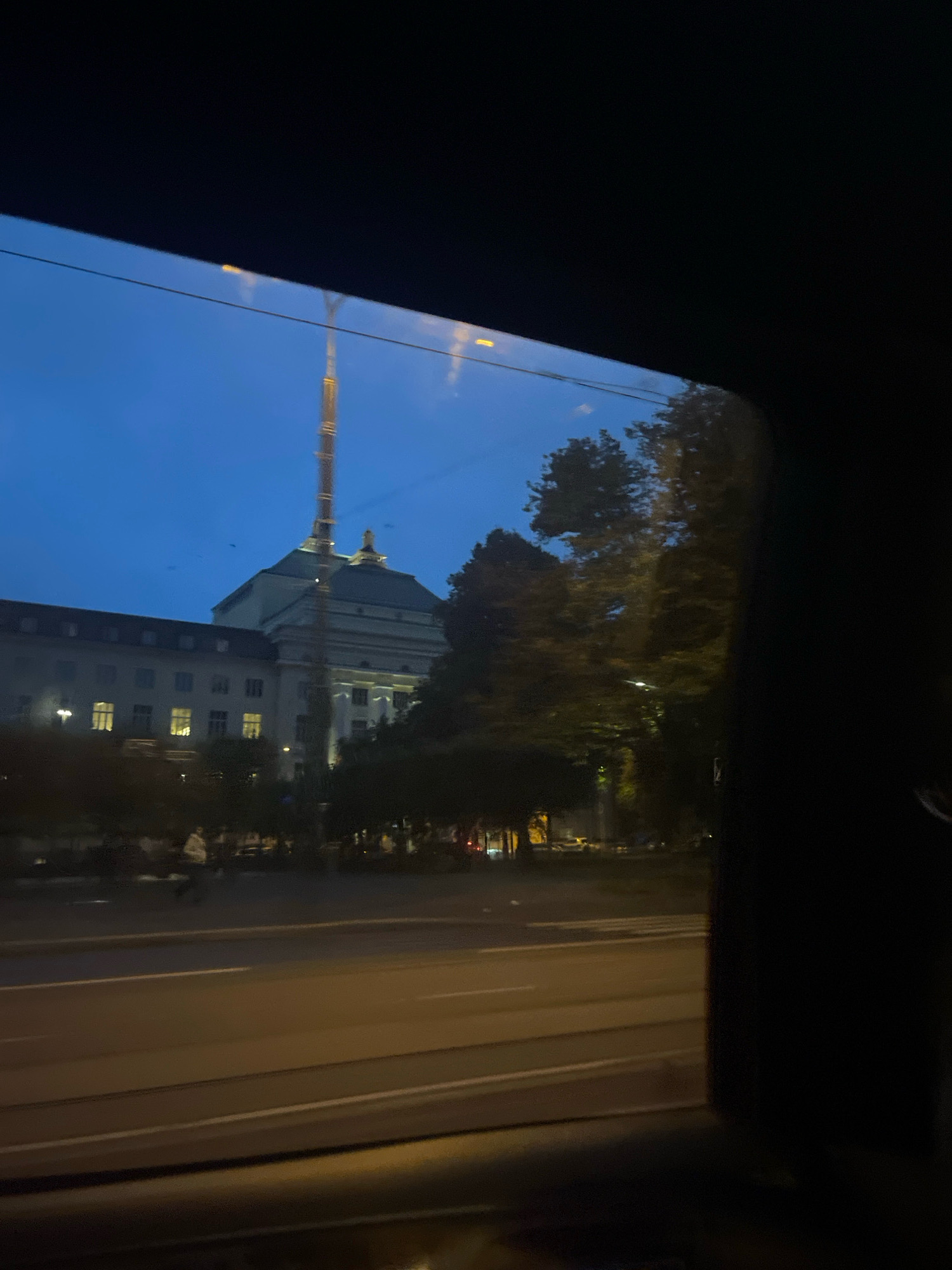 This is a nighttime, low-angle, medium shot captured from inside a vehicle, looking out a window at a building that appears to be the Estonian Academy of Arts in Tallinn, Estonia. The building is well-lit with warm interior lights visible through its windows, and its grand architecture, including a domed roof and a tall spire, is prominent. Trees with dark foliage frame the right side of the building. In the foreground, a street with multiple lanes and what appears to be tram tracks is visible, with faint streetlights illuminating the scene. A few blurry figures are seen walking on the sidewalk and crossing the street. The sky is a deep twilight blue, with a few dark birds visible against it. The image is partially obscured by the dark interior of the vehicle, creating a framing effect.