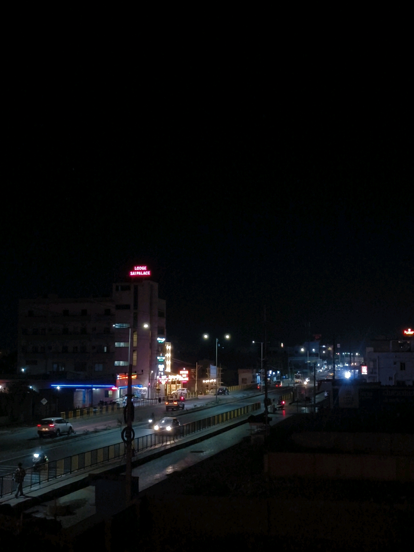 The image depicts a nighttime scene in Vijayapura, India, likely on a busy road or highway. A multi-story building with a neon sign that reads "LODGE SAI PALACE" is visible in the upper left portion of the frame, illuminated against the dark sky. Cars are driving on a road below, their headlights and taillights creating streaks of light. There are streetlights illuminating the road and sidewalk. A pedestrian is visible walking on a sidewalk in the lower left part of the image. The overall atmosphere is one of urban activity at night, with artificial lights dominating the scene.