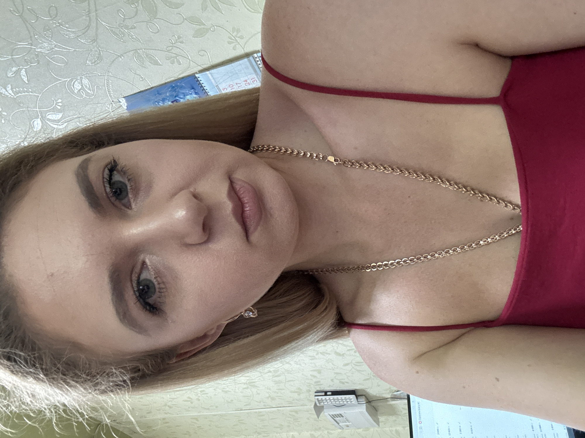 A blonde woman with a gold chain necklace and a red tank top is shown in a medium close-up shot. She has light skin and her hair is pulled back, with some strands falling around her face. The background appears to be an indoor setting with patterned wallpaper and a calendar visible. There is a computer monitor to the left of the frame.