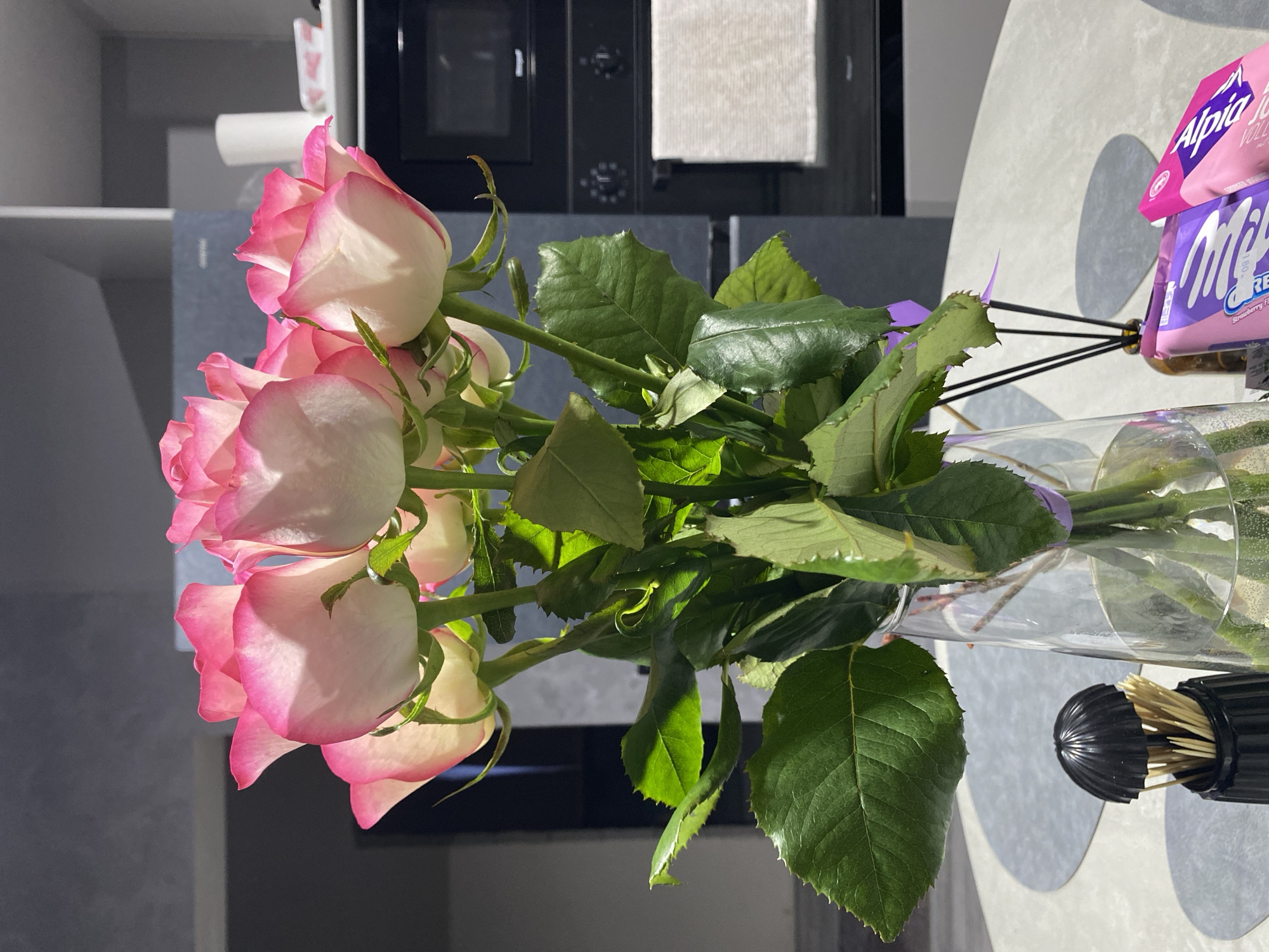 A vase of pink and white roses sits on a table in an indoor environment. The roses are in full bloom with bright green leaves. The vase is transparent, revealing the stems. Next to the vase is a black container with wooden skewers. In the background are kitchen appliances, including a microwave and a storage cabinet. The lighting suggests a natural light source. The image was taken in the city of Industrialny, Russia. There are packs of candy next to the vase.