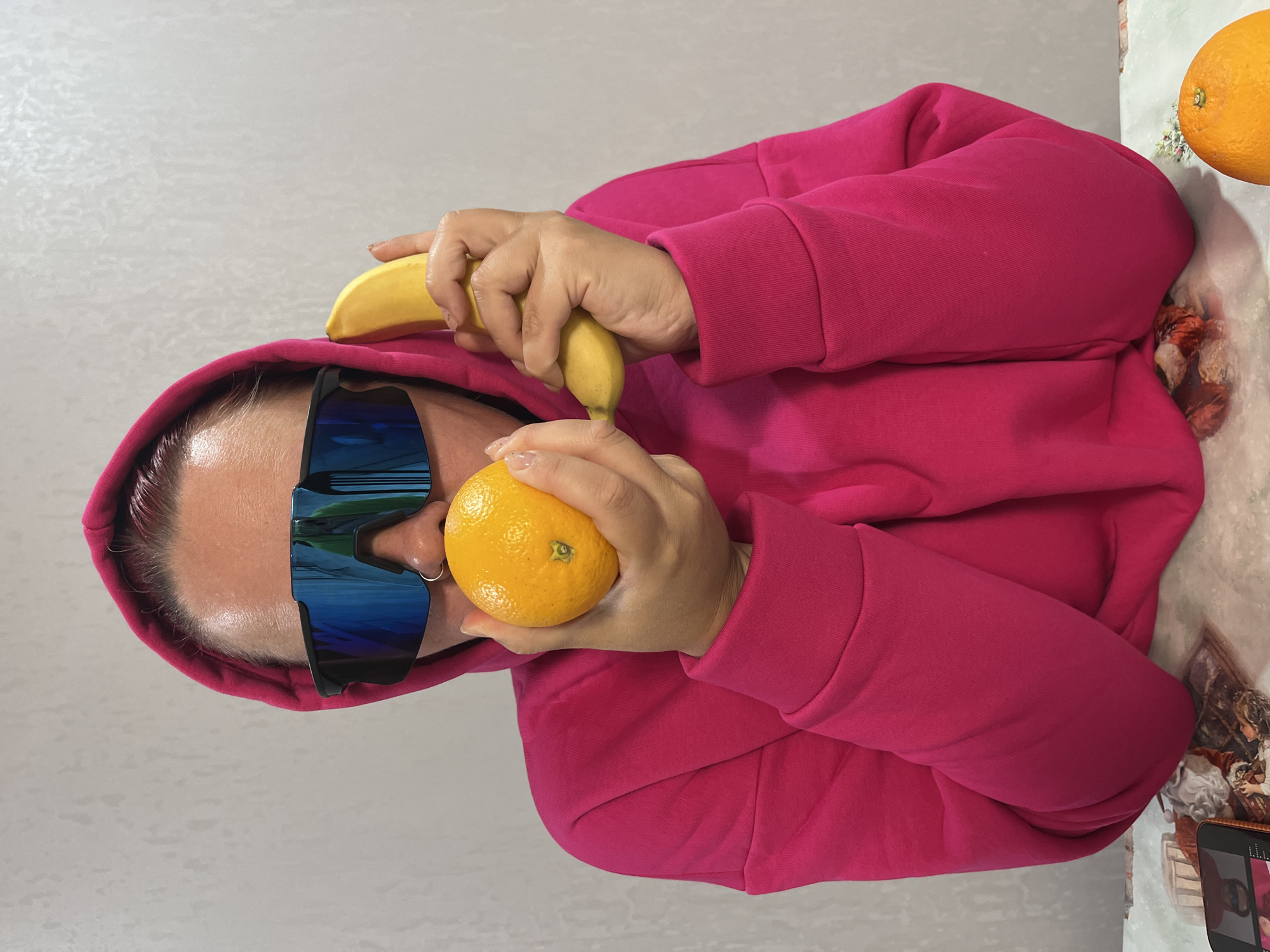 The image depicts a person wearing a bright pink hooded sweatshirt and large, mirrored sunglasses. The person is holding a banana in their left hand and an orange in their right hand, positioning the orange in front of their mouth. Part of another orange is visible in the upper right corner of the image. The background is a plain, light-colored wall, and the surface below the person is covered with a decorative tablecloth. A smartphone is visible in the lower right corner. The location is not evident from the provided image. There is nothing in the image that indicates it is located in the city of Chaikovsky, Russia.