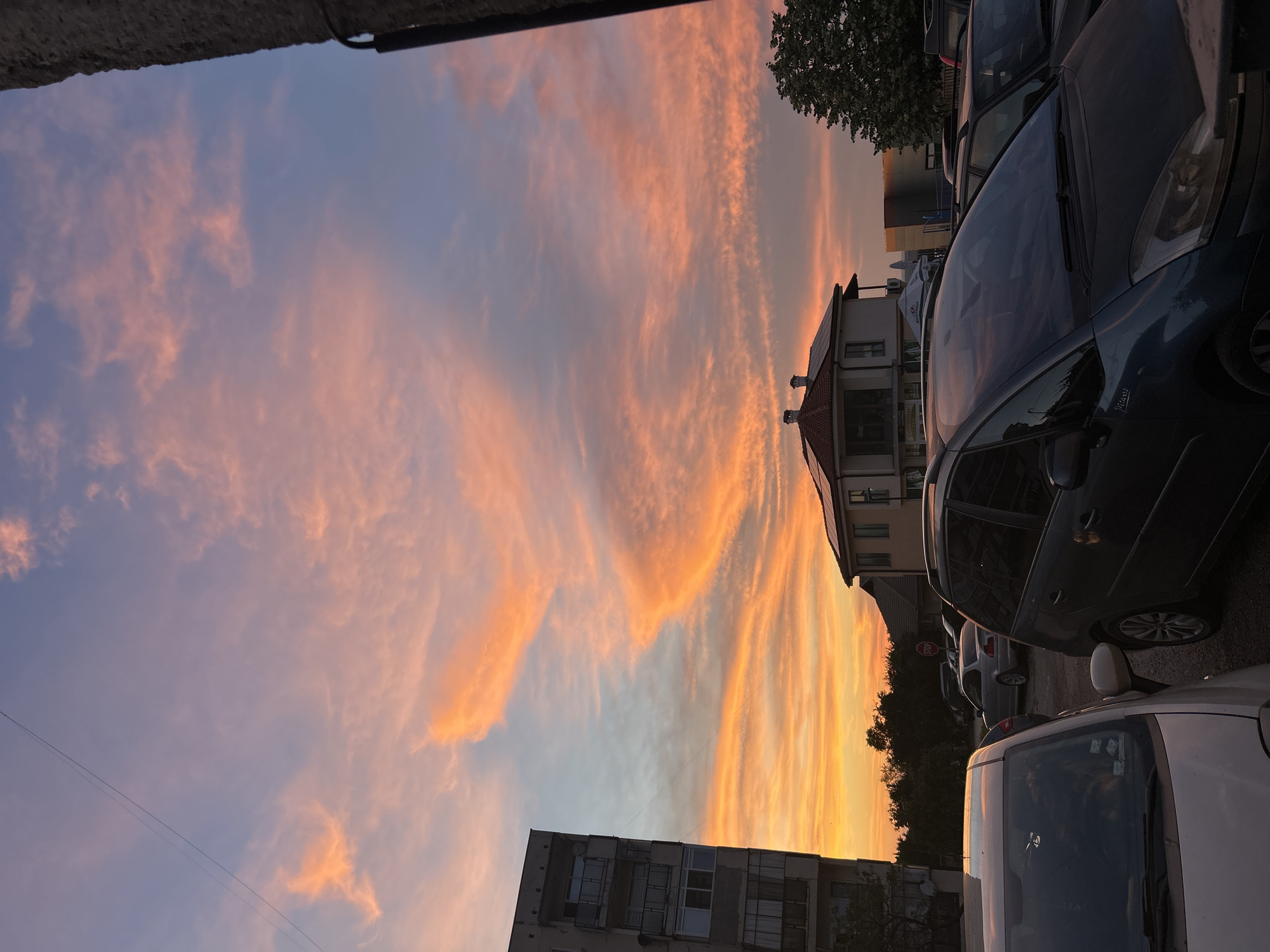 This is a medium shot, eye-level photograph taken during sunset. The sky is filled with colorful clouds ranging from orange to pink and blue. In the foreground, several cars are parked. A dark blue minivan is prominently in the center, with its windshield reflecting the sky. To its left is a lighter-colored car, and behind it, other vehicles are visible. On the left side of the image, a tall, grey apartment building stands, with balconies and windows. In the middle ground, a house with a red roof and solar panels is visible, surrounded by trees. Further to the right, another building and a tree are present. A stop sign is also visible among the parked cars. The overall scene depicts an urban setting bathed in the warm glow of a sunset.