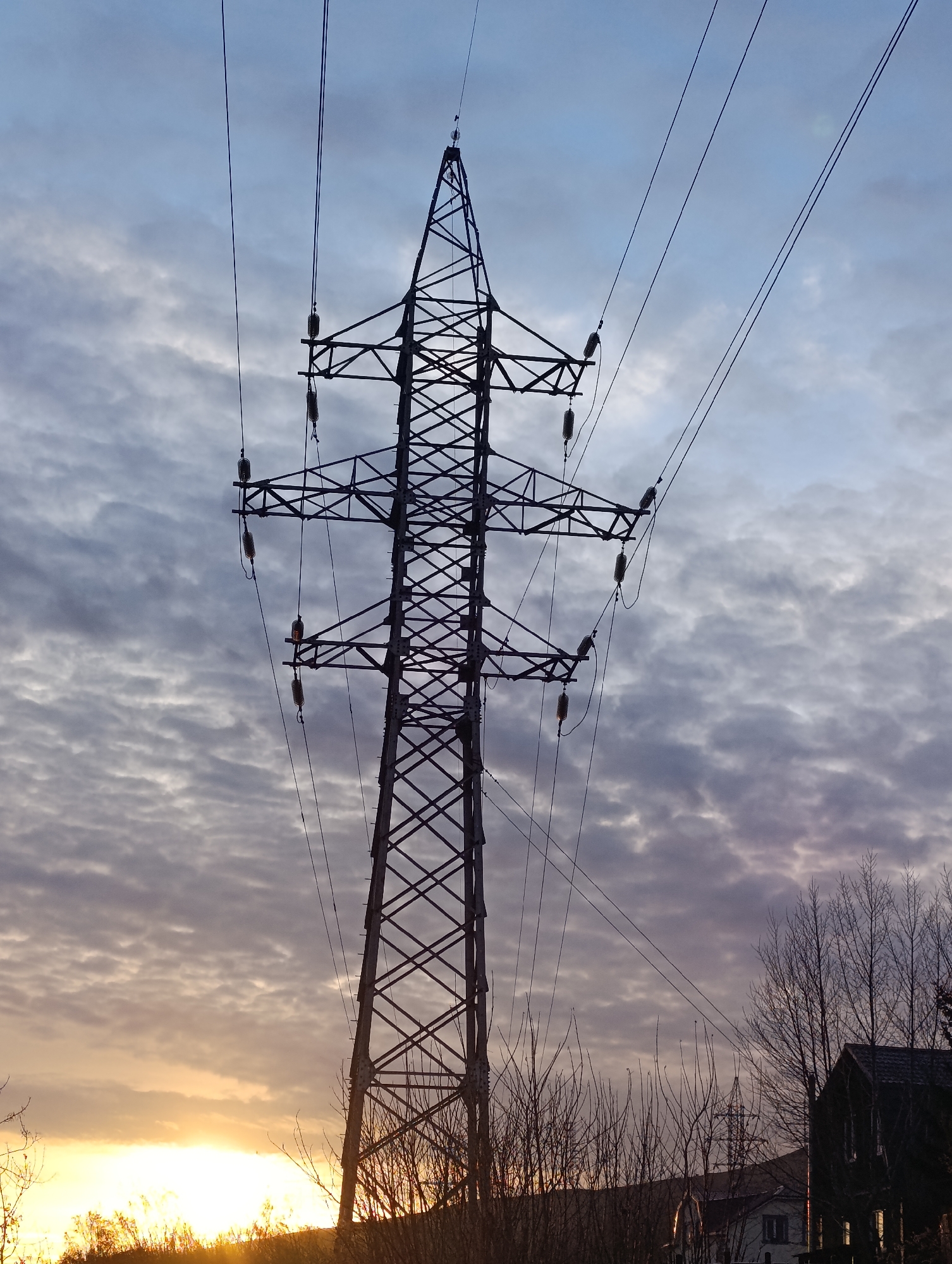 The image features a steel lattice transmission tower standing prominently against a sky filled with textured clouds at sunset. The tower is equipped with insulators and conductors, and is placed in an area with some trees and low vegetation at the base. In the lower right, parts of houses can be seen, suggesting an inhabited area. Light from the setting sun illuminates the base of the tower and surrounding vegetation. Location information suggests this image was taken in Krasnoyarsk, Russia.