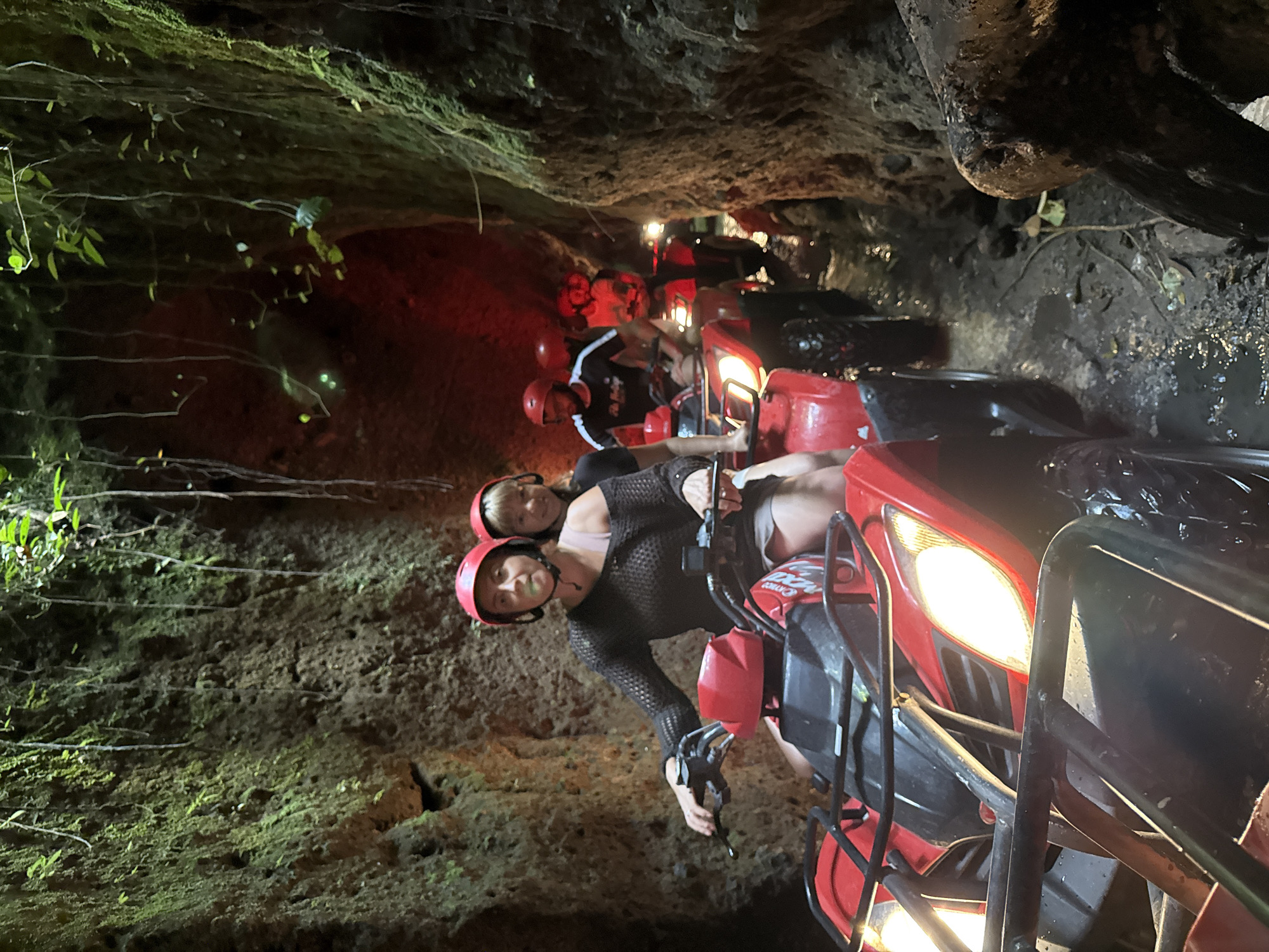 An All-Terrain Vehicle (ATV) tour is depicted within a narrow, natural passage in Payangan, Indonesia. The scene shows a line of individuals driving red ATVs through a confined, wet, and muddy gorge.
In the immediate foreground, a woman wearing a red helmet and a black open-knit long-sleeved top over a light-colored inner garment operates a red ATV with black trim and exposed metal racks. Her hands are on the handlebars, and she appears focused forward. Behind her, partially visible, is another woman with blonde hair, also wearing a red helmet, seated as a passenger. The ATV's bright white headlights are illuminated, casting light on the muddy ground and the textured walls.
Behind the lead ATV, a queue of at least three additional red ATVs and their riders proceeds through the passage. One male rider in the mid-ground wears a red helmet and a black short-sleeved shirt with white stripes on the sleeves. The headlights of the trailing ATVs are also illuminated, and a strong reddish glow emanates from further down the passage, illuminating the distant walls in red.
The passage walls are irregular and high, composed of natural rock or compacted earth, densely covered with green moss, small plants, and numerous hanging vines, particularly on the left side of the frame. The ground is consistently muddy and wet, with visible puddles and tire tracks from the ATVs. The overall environment is dim, with artificial light from the ATV headlights being the primary illumination source within the dark, enclosed space.