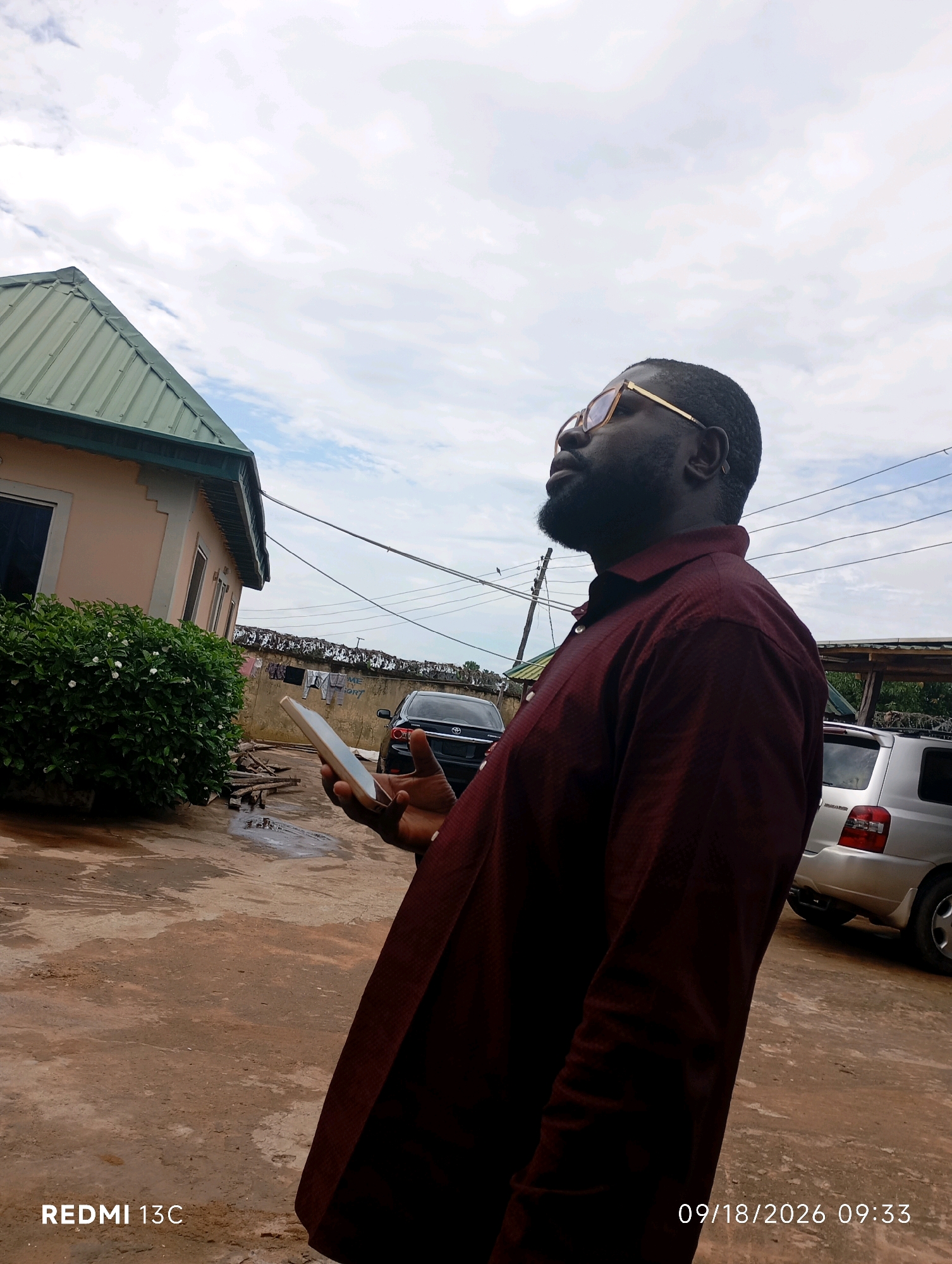 The image depicts an outdoor scene, likely in the city of Shendam, Nigeria, on September 18, 2026, at 9:33 AM. A man with dark skin, wearing glasses and a dark red collared shirt, is positioned in the foreground, looking upwards and to the right. He is holding a white smartphone in his left hand. Behind him, a light pink building with a green corrugated metal roof is visible, along with a dense green shrubbery in front of it. Further in the background, a black car and a silver SUV are parked. The ground appears to be dirt or concrete, with some puddles of water. Power lines and a wall with laundry hanging on it are also present, indicating a residential or semi-urban setting. The sky is overcast with clouds.
