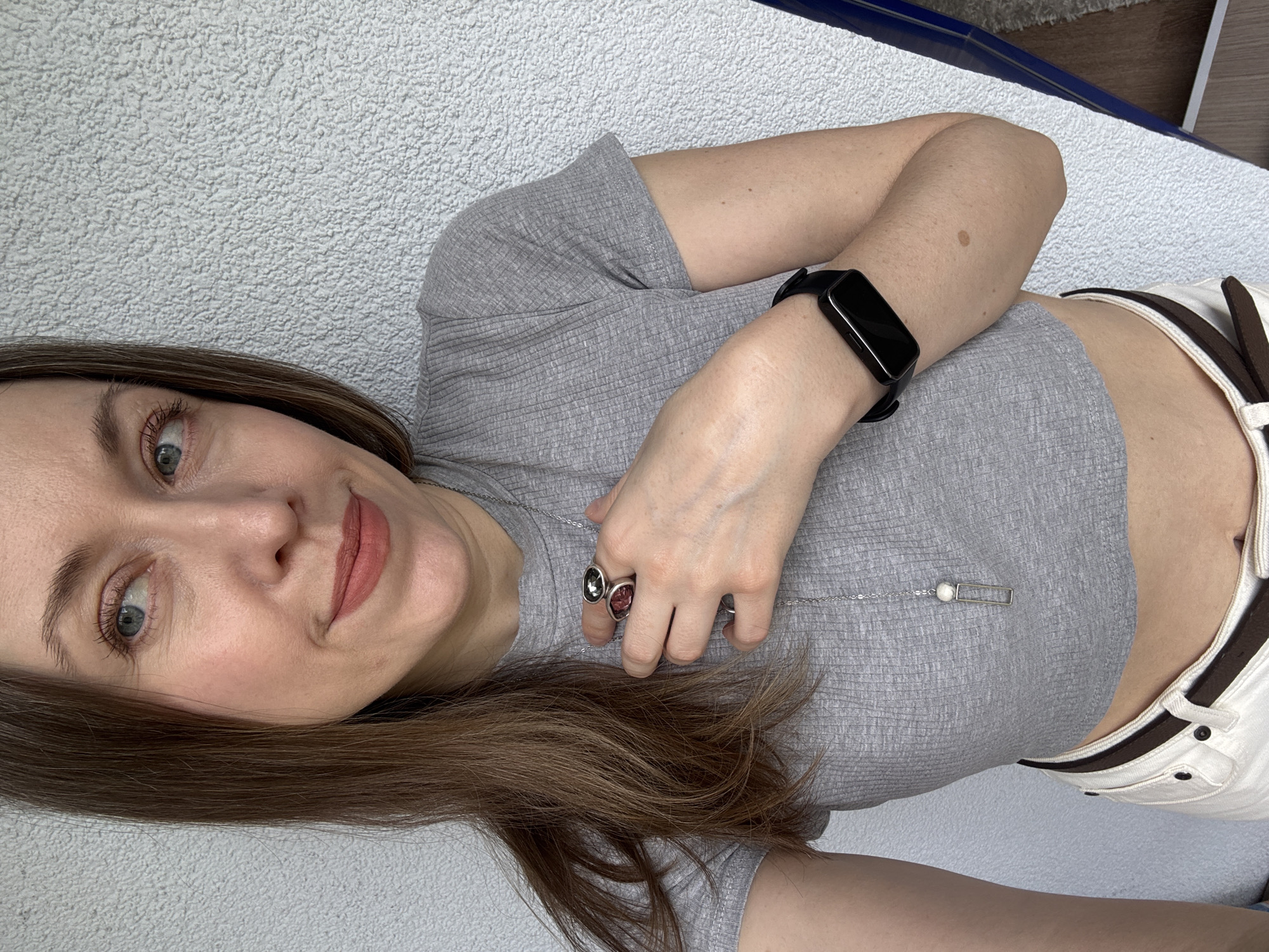 A woman lies on a light gray textured surface, likely fabric, with part of a white and brown garment visible on the right side of the frame. She is wearing a short-sleeved gray ribbed top, a black smartwatch on her left wrist, and two rings on her left hand, one with a gray stone and another with a pink stone. She also has a silver necklace. She is looking towards the camera with a slight smile. Her hair is brown and shoulder-length. There is no explicit indication of Kudrovo, Russia, in the image.